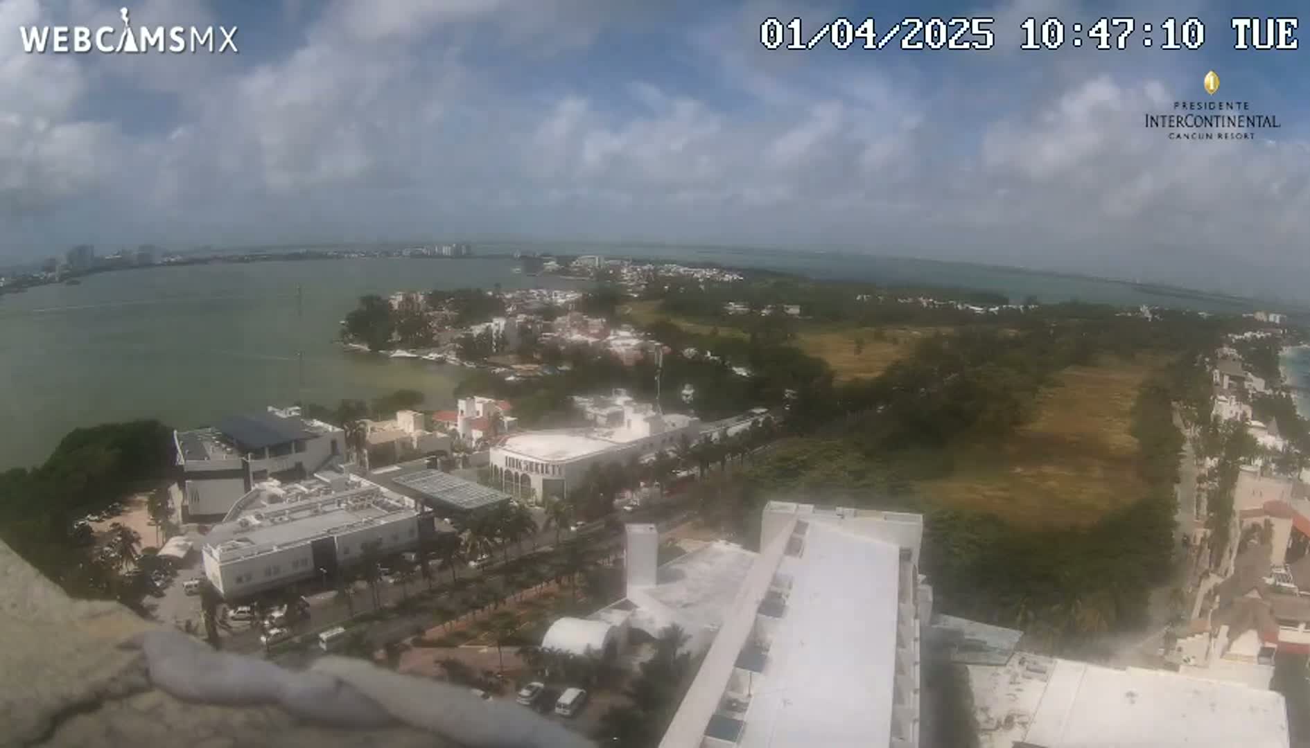 A high-angle, outdoor view on a partly cloudy day shows a coastal city with buildings, vegetation, and a body of water in the background.