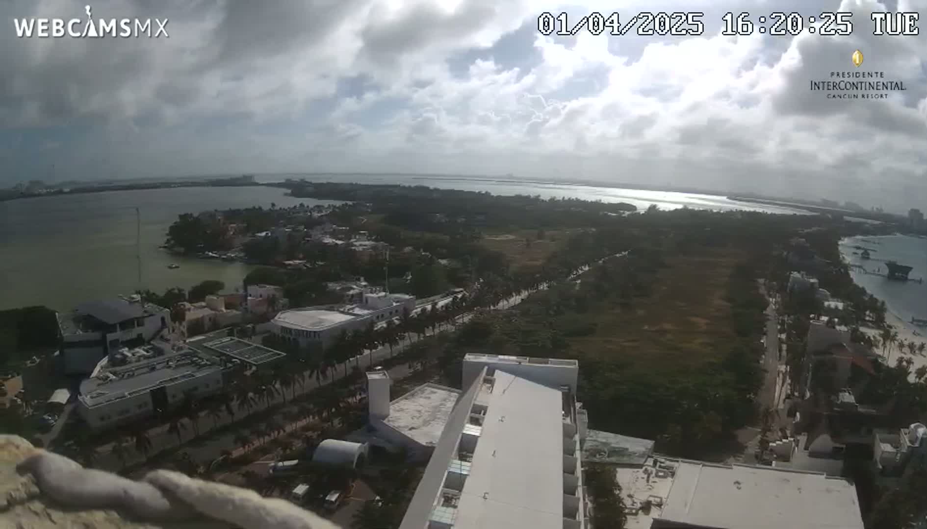 A high-angle view of a coastal city on a partly cloudy day shows a body of water, buildings, and vegetation, with a section of a large building in the foreground.