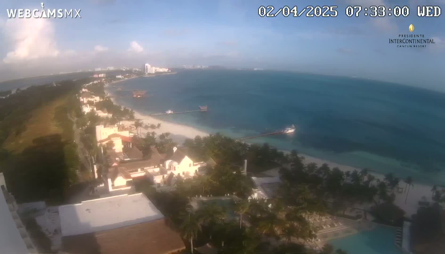 An aerial view of a tropical coast shows a beach with palm trees, buildings, and calm ocean water under a mostly clear sky.