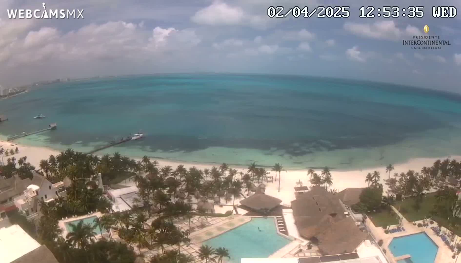 An aerial view shows a beach resort with swimming pools next to a white sand beach and turquoise ocean under a partly cloudy sky.