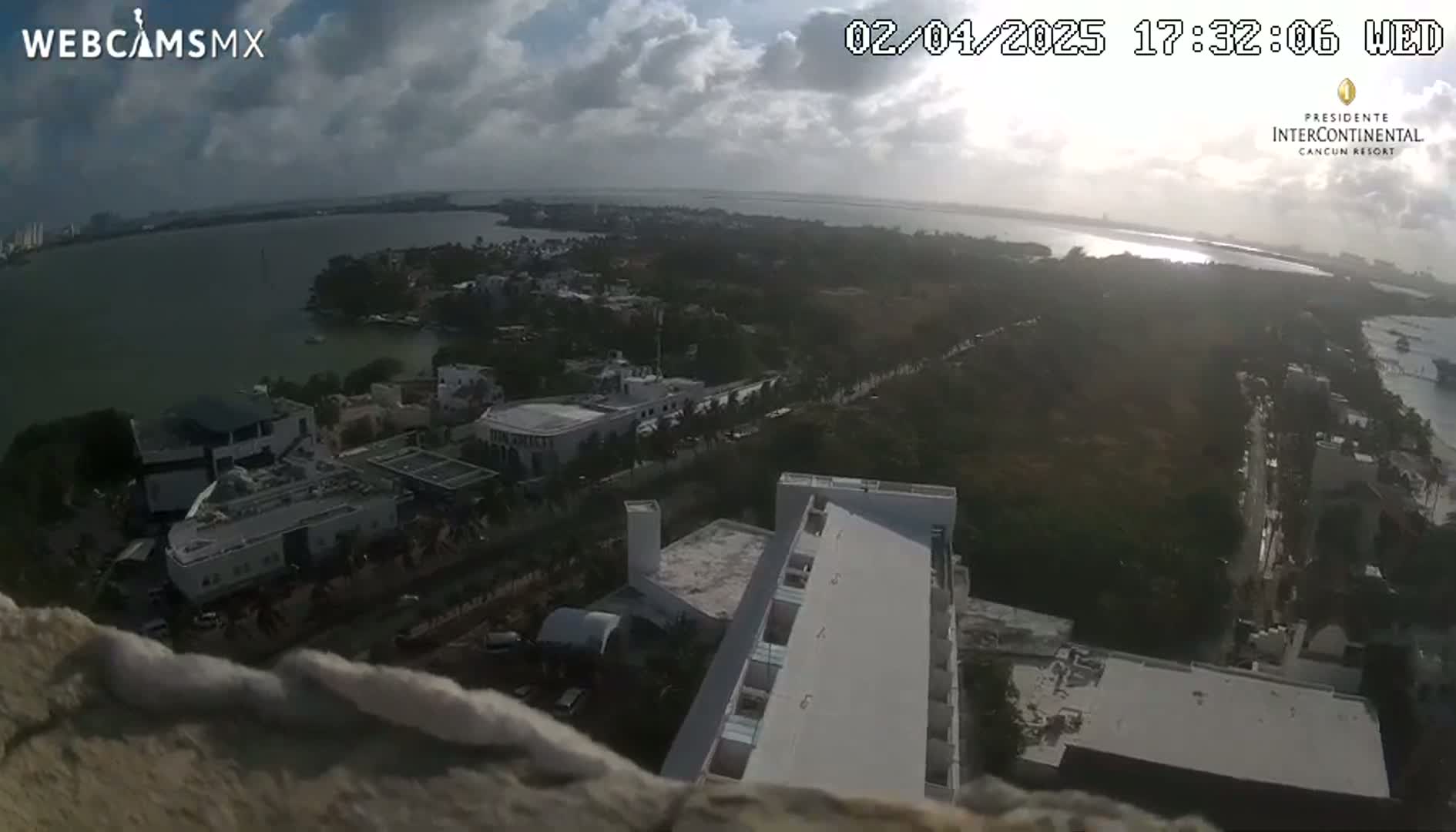 An aerial view shows a partly cloudy day overlooking a body of water, buildings, and lush vegetation.