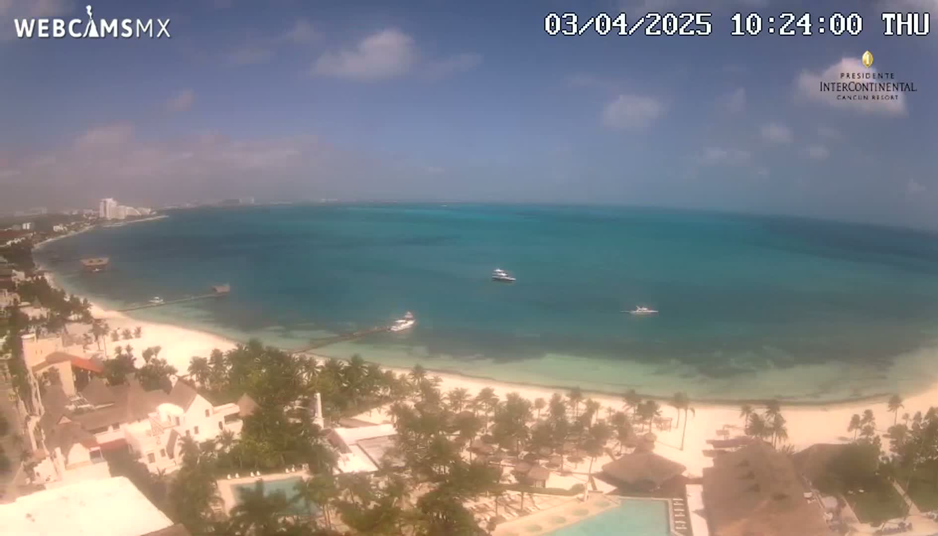 An aerial view shows a tropical beach with clear, shallow turquoise water, a few boats, and palm trees under a partly cloudy sky.