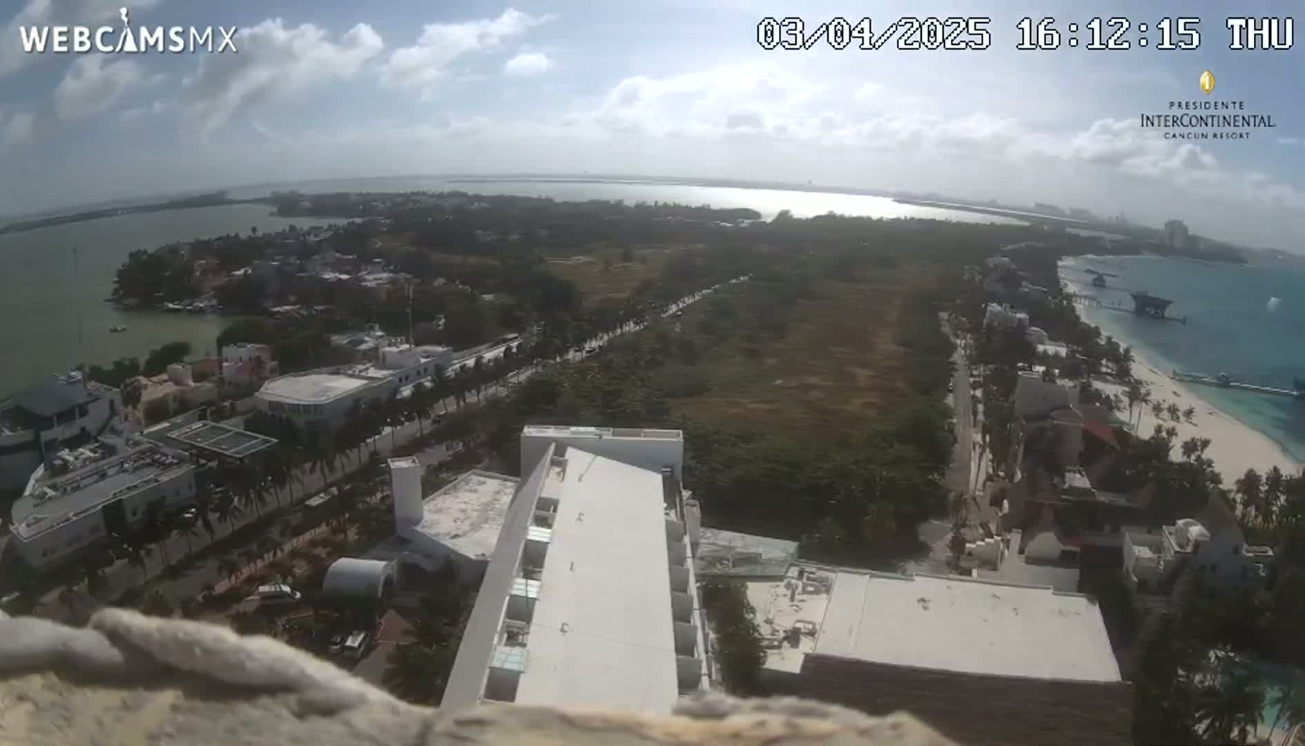 A high-angle, sunny view shows a coastal city with buildings, roads, and vegetation near a body of water under partly cloudy skies.