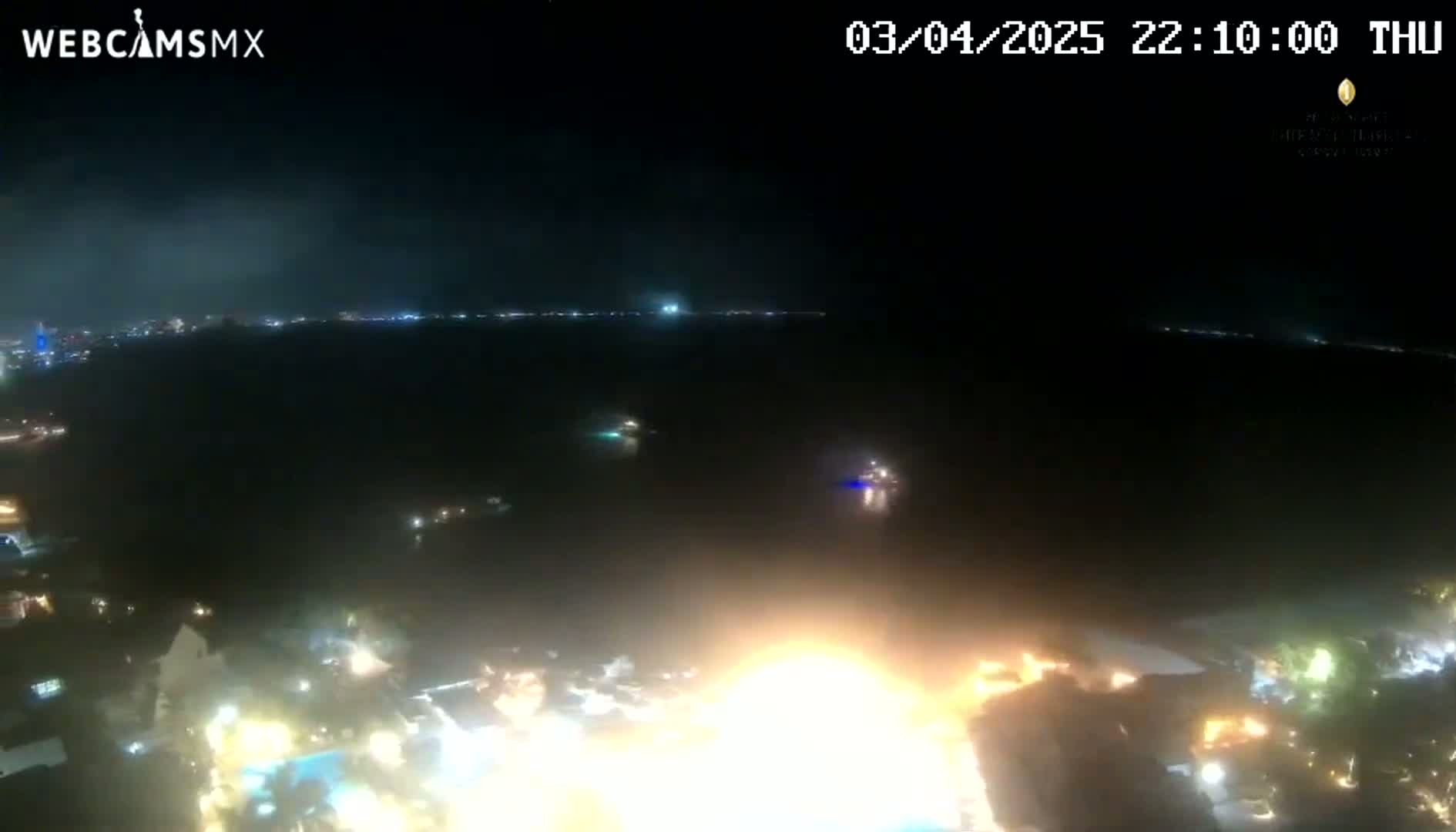 A nighttime aerial view shows a body of water with lights on the far shore and a brightly lit area closer to the camera with a large fire or intense light source.