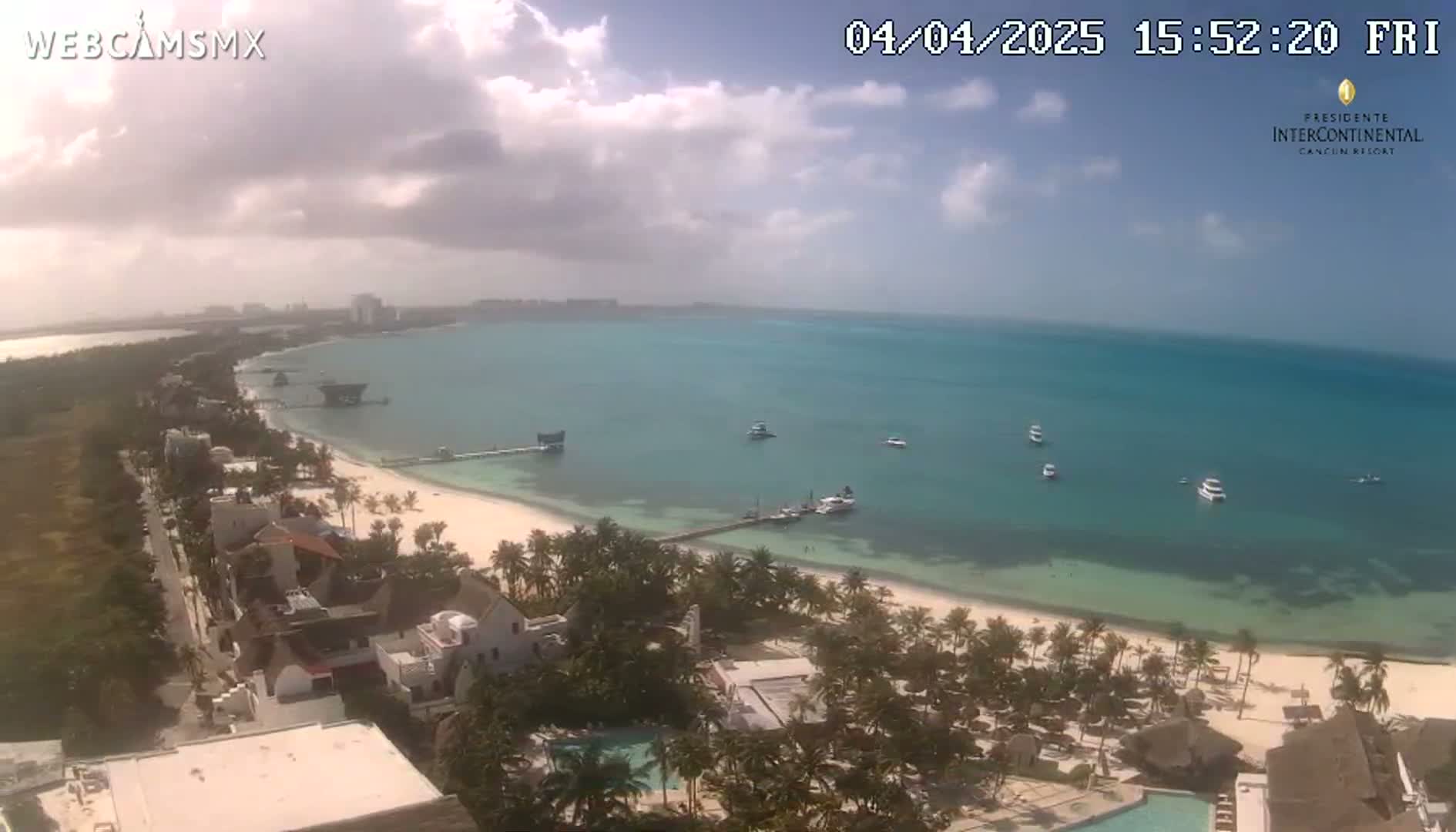 An aerial view shows a partly cloudy day at a beach with turquoise water, white sand, palm trees, and several boats offshore.