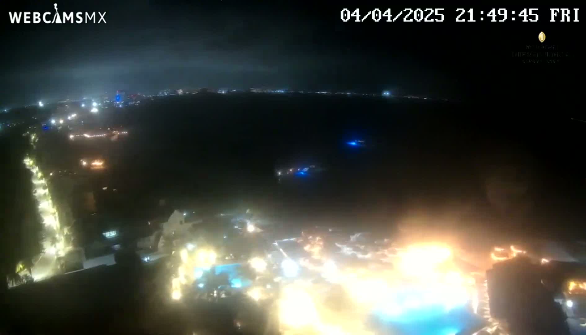 A nighttime aerial view shows a brightly lit resort area with a large pool and surrounding buildings, under a mostly clear, dark sky.
