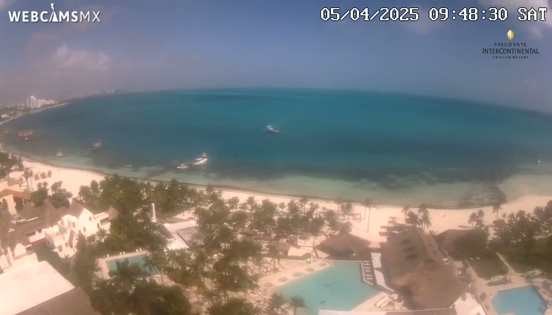 An aerial view shows a beach with turquoise water, a resort with swimming pools, and palm trees under a mostly clear sky.