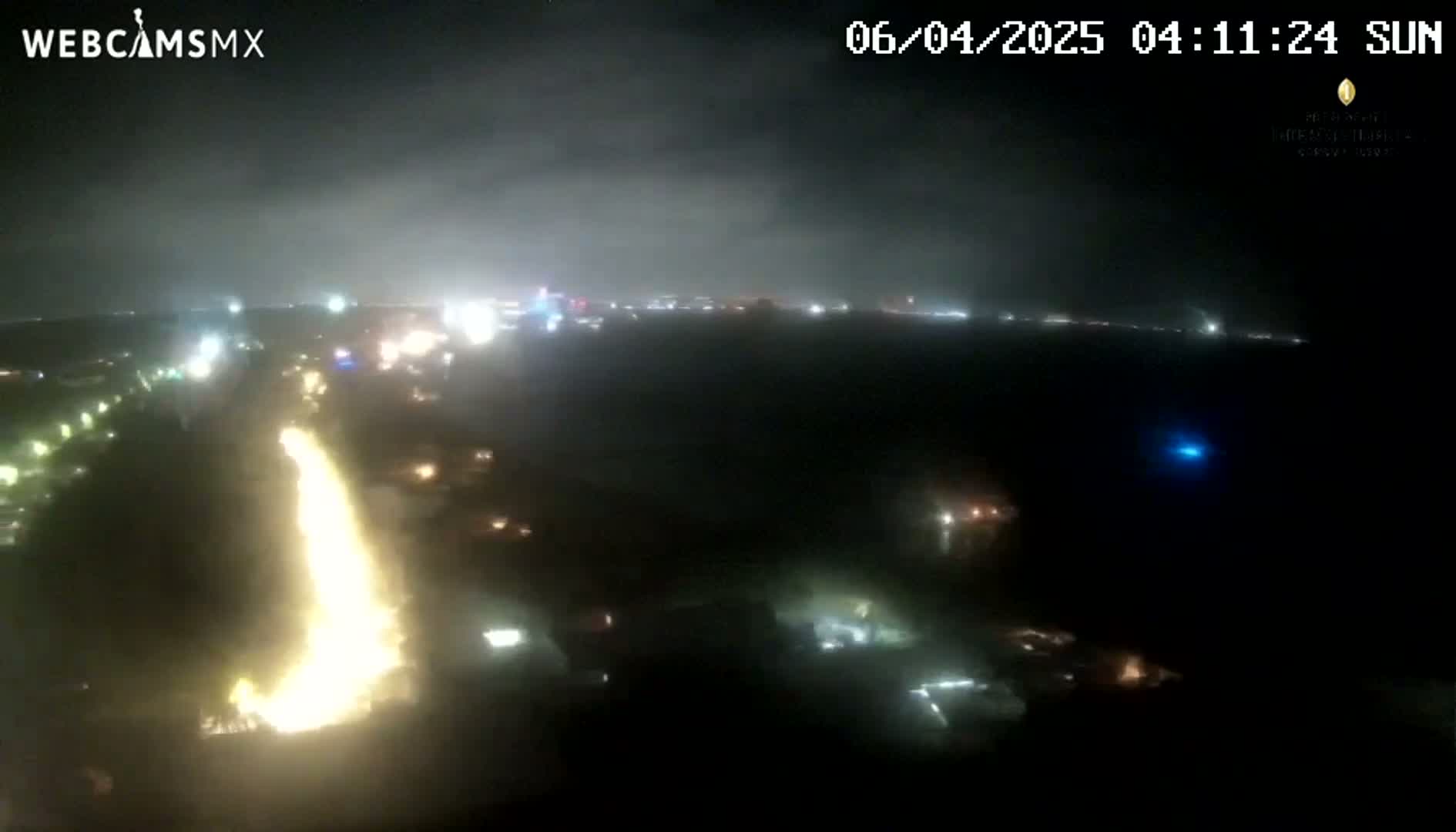 A nighttime aerial view shows a bright, vertical, yellowish light source amidst scattered city lights under a cloudy sky.