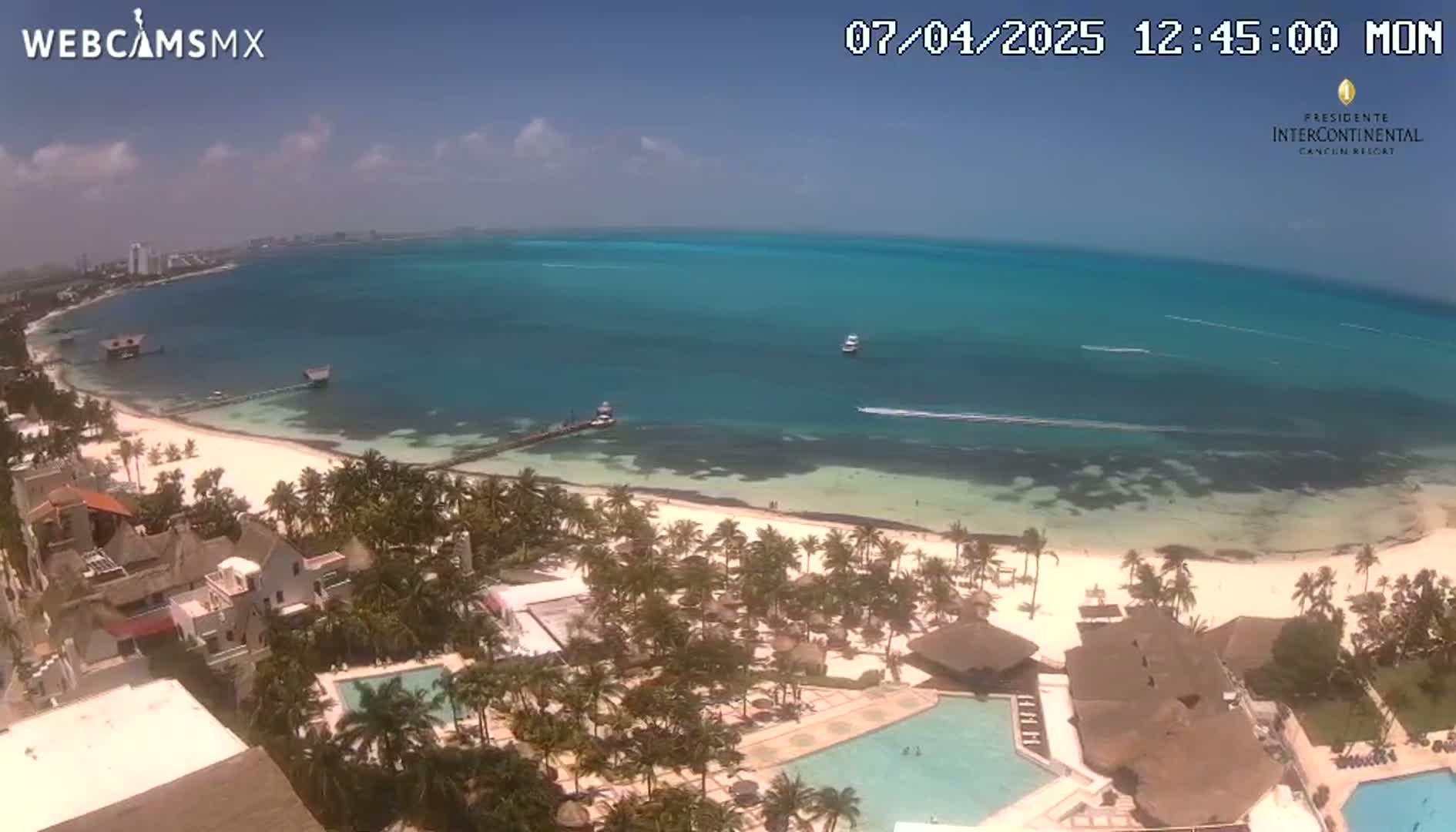 An aerial view shows a tropical beach with clear turquoise water, palm trees, and a resort with swimming pools under a sunny sky.