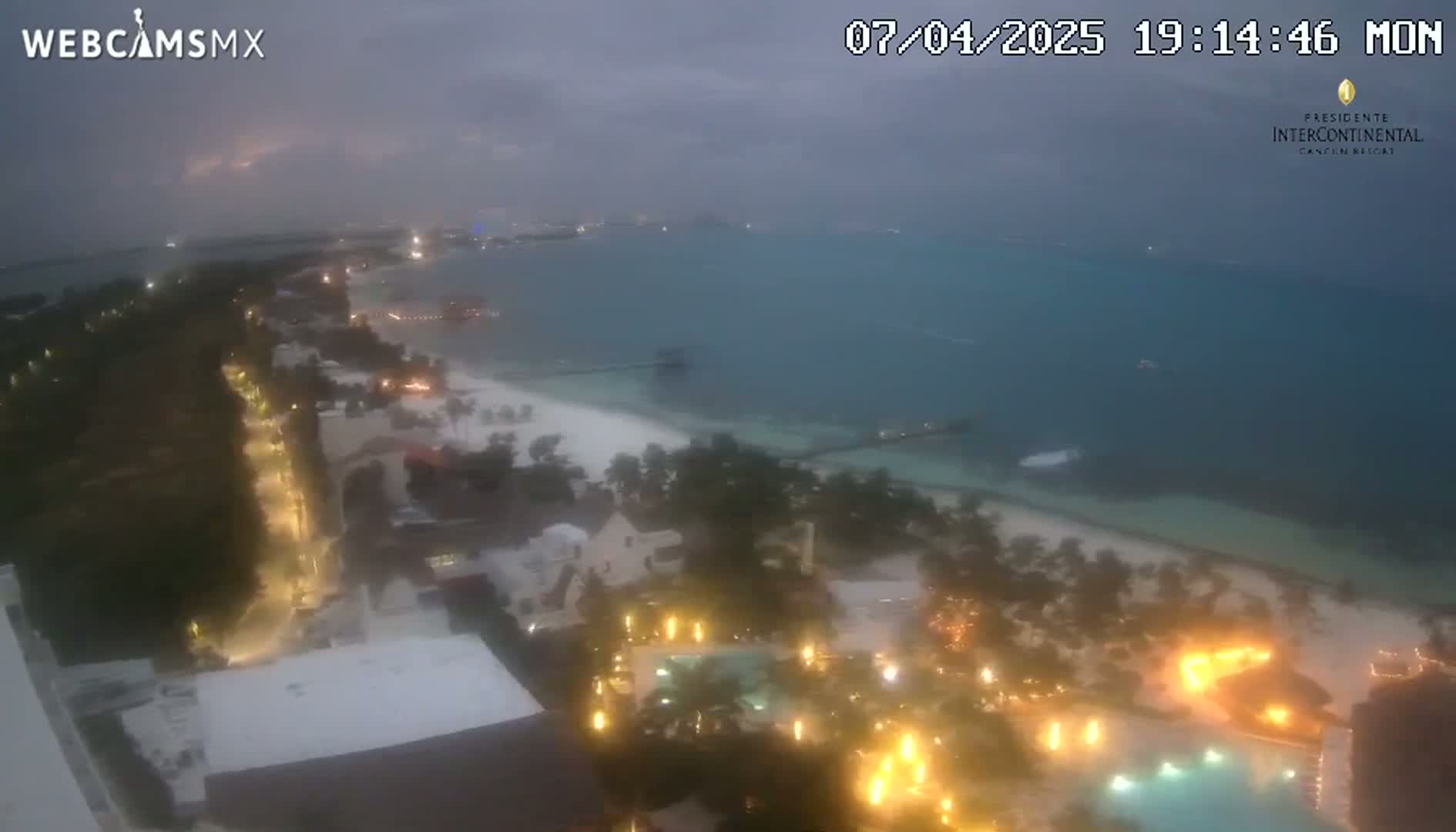 An aerial nighttime view of a beach resort area shows a mostly cloudy sky over a calm ocean with a sandy beach, illuminated buildings, and pools.