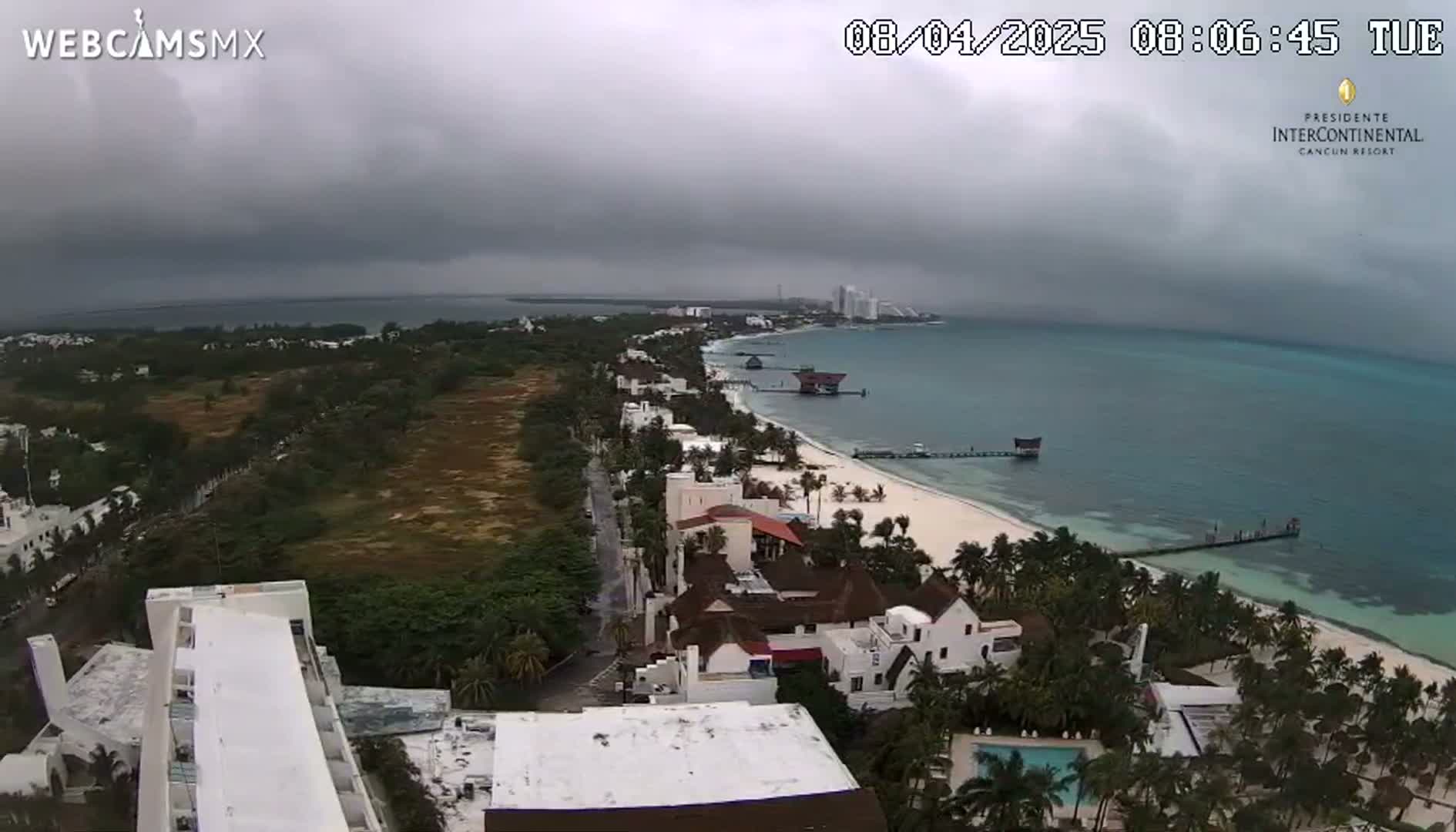 An aerial view of a coastal city under an overcast sky shows a white sand beach, turquoise water, buildings, palm trees, and a road.