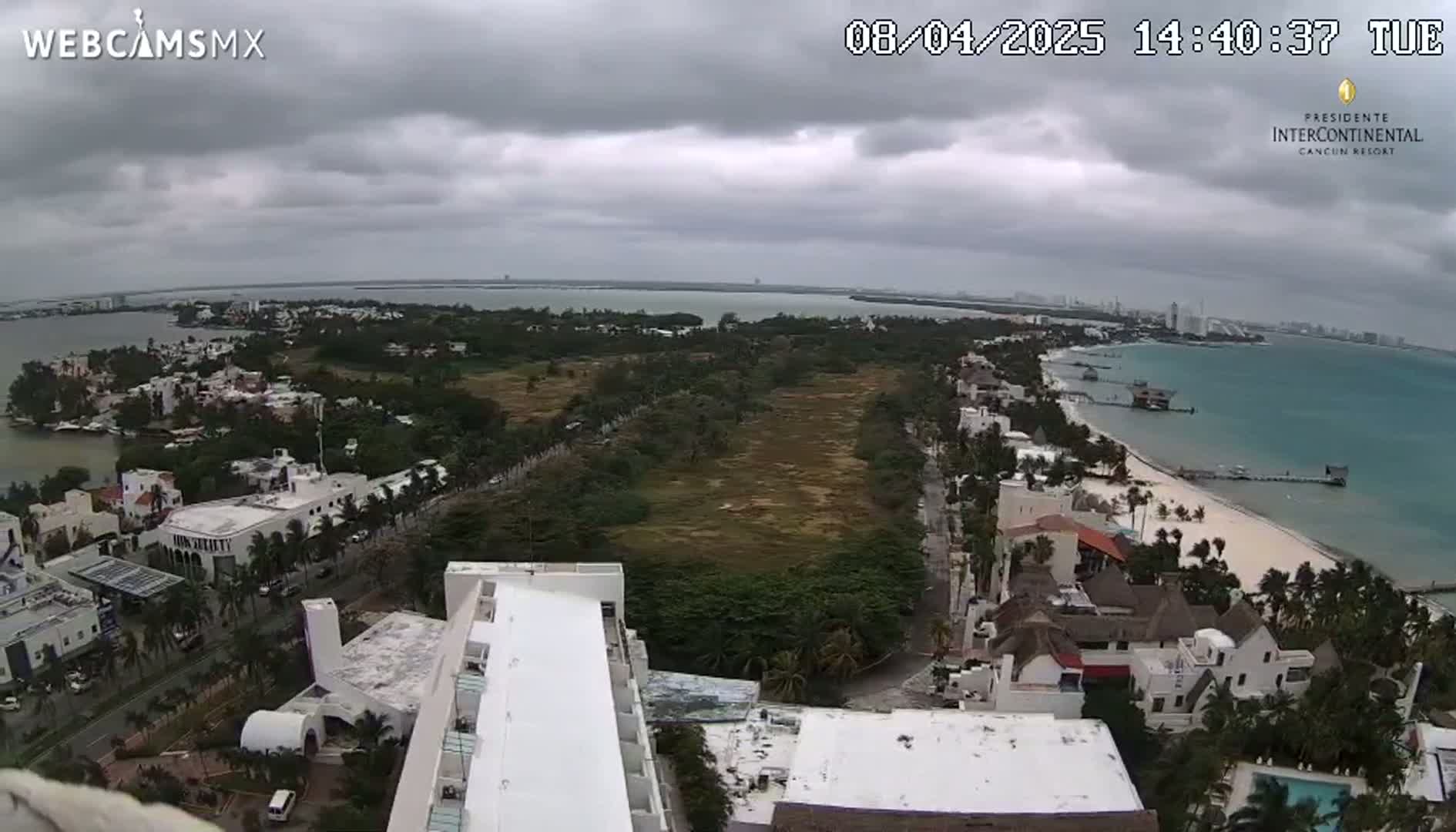 An elevated view of a coastal town on an overcast day shows a sandy beach, turquoise water, and buildings, with a large undeveloped area separating the beach from the buildings inland.