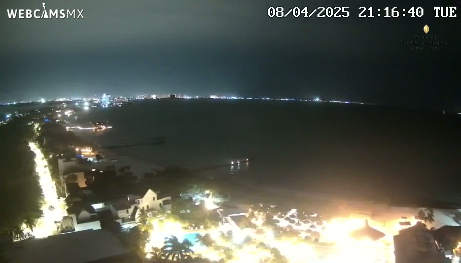 A nighttime, high-angle view shows a city skyline across a dark body of water, with brightly lit beachfront properties and palm trees in the foreground.