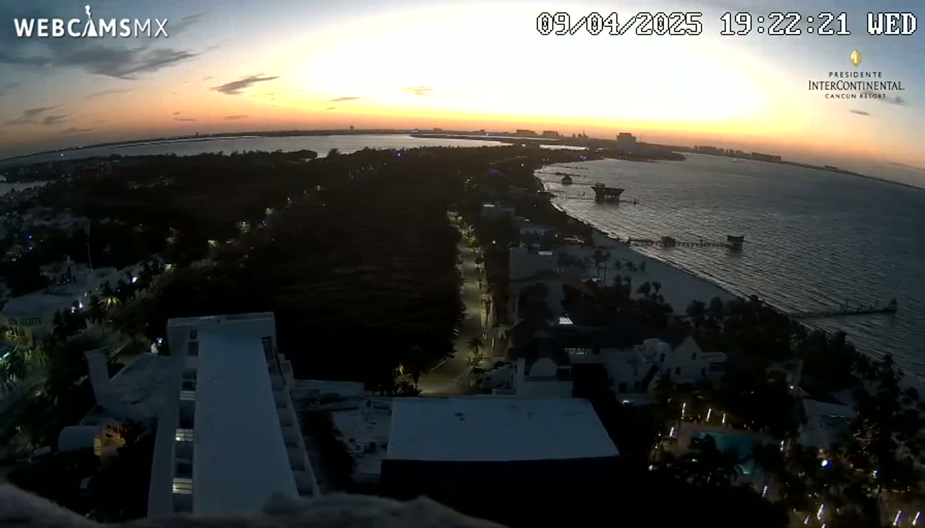 An aerial view at sunset shows a coastal resort town with a beach, ocean, and buildings, illuminated by lights as the sun sets over calm water.