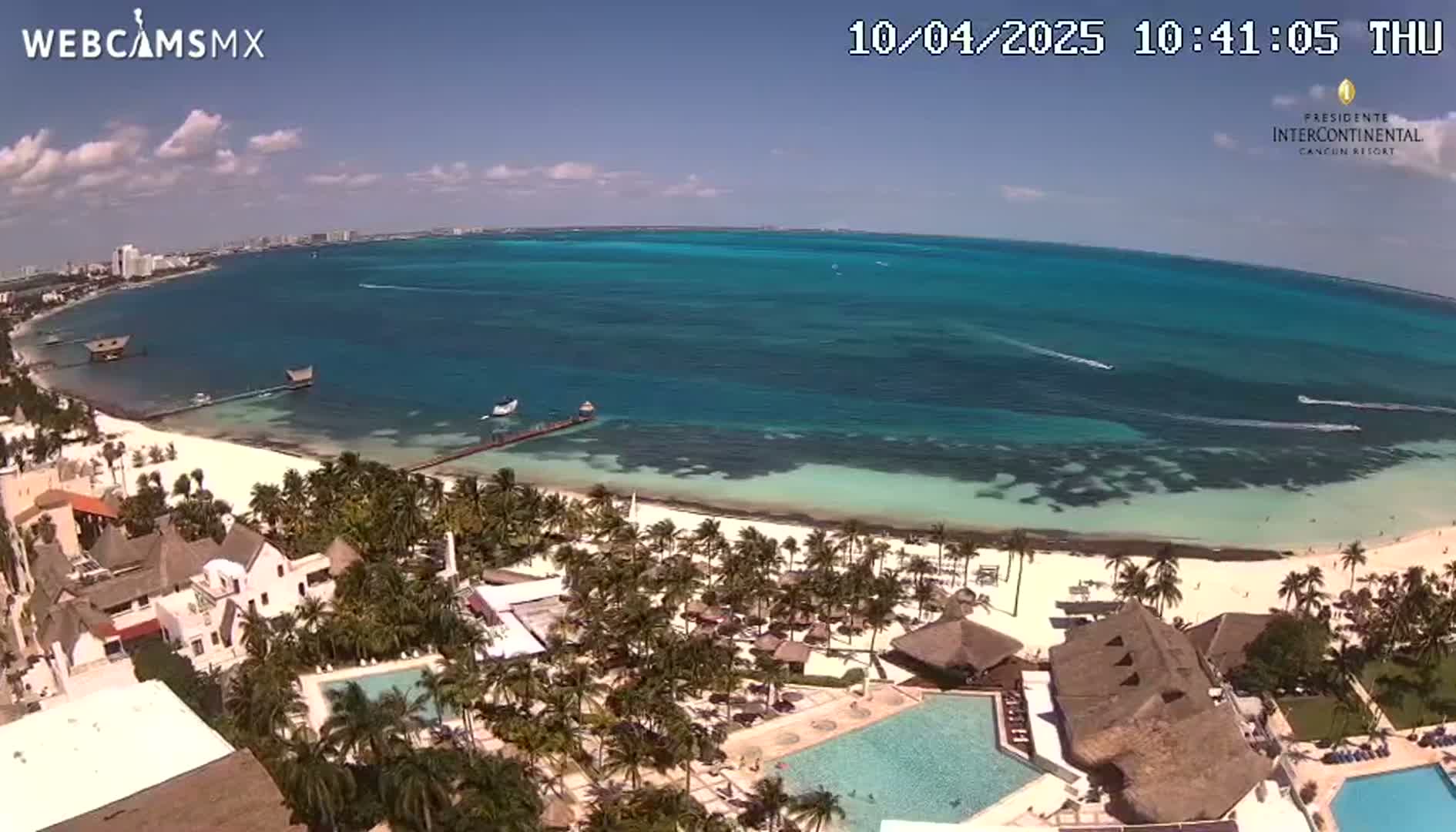 An aerial view shows a resort on a white sand beach with turquoise water under a mostly sunny sky.