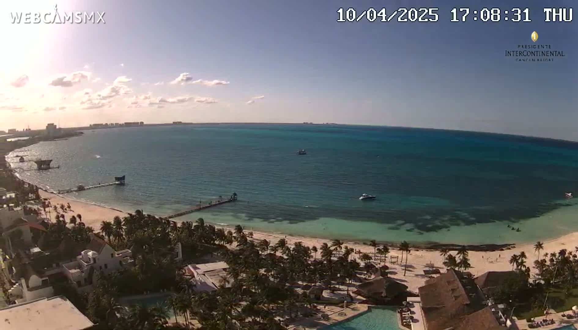 An aerial view shows a sandy beach, turquoise water, palm trees, and resort buildings under a mostly sunny sky.