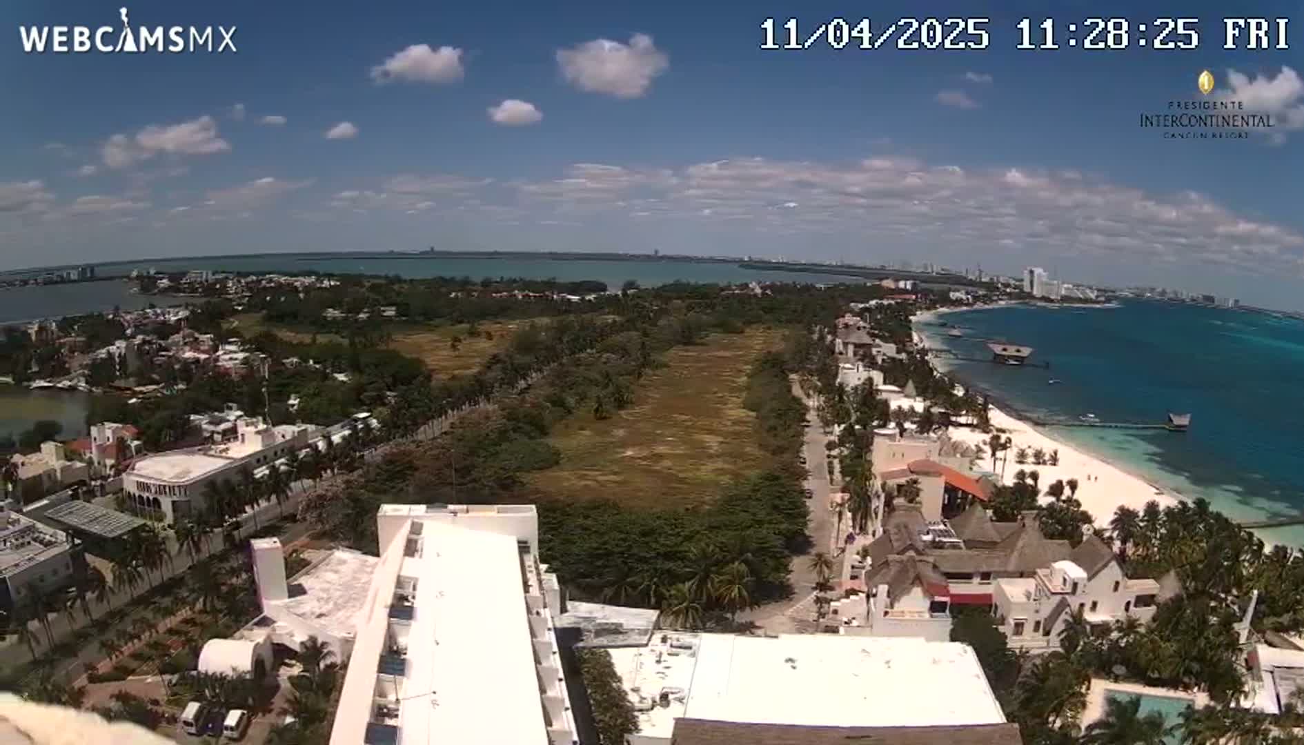 An aerial view of a coastal town on a partly cloudy day, showing buildings, vegetation, a beach, and calm ocean water.