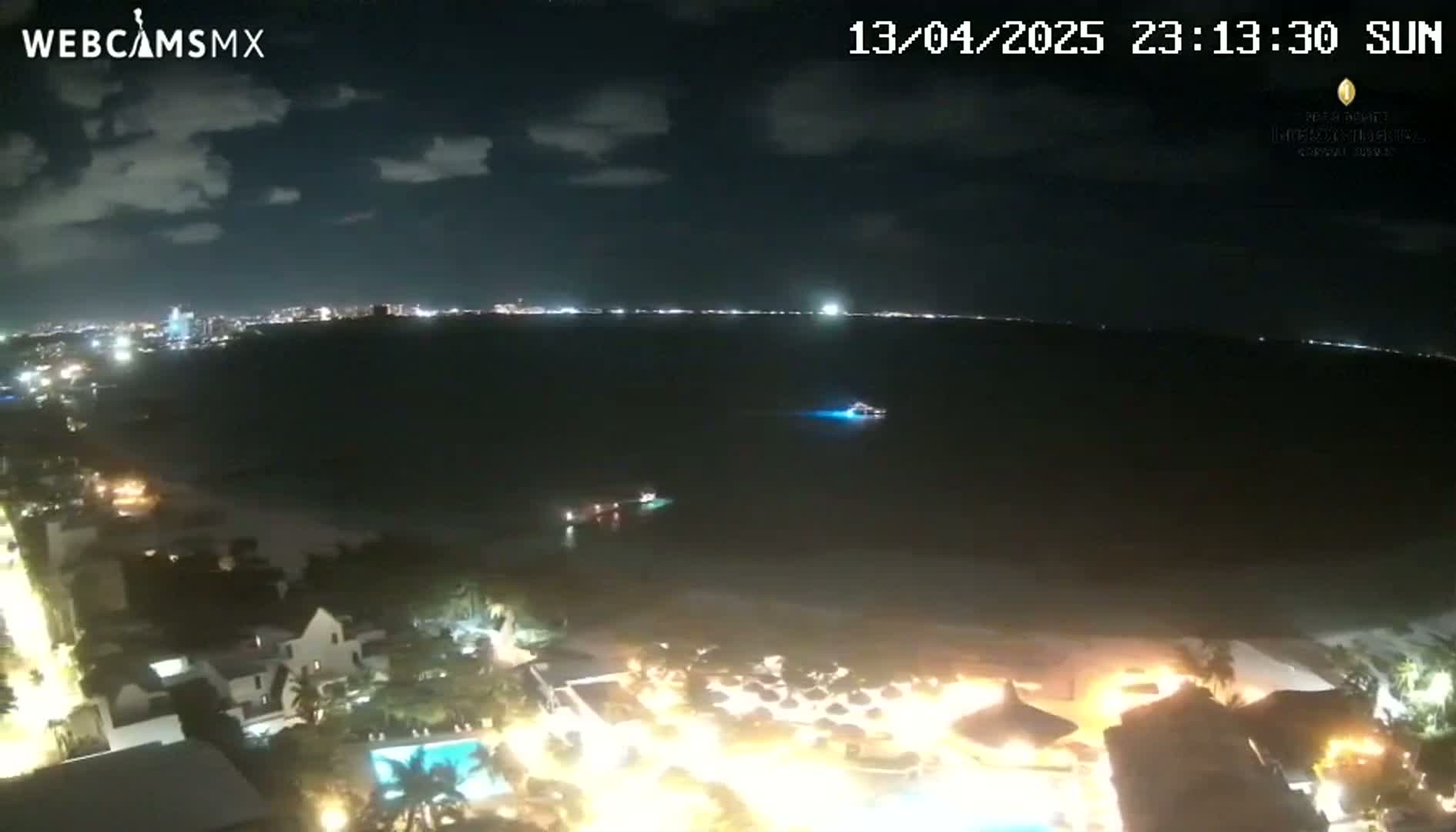 A nighttime coastal scene shows a dark body of water with a few boats, a brightly lit shoreline with buildings and pools, and a city skyline in the distance under a partly cloudy sky.