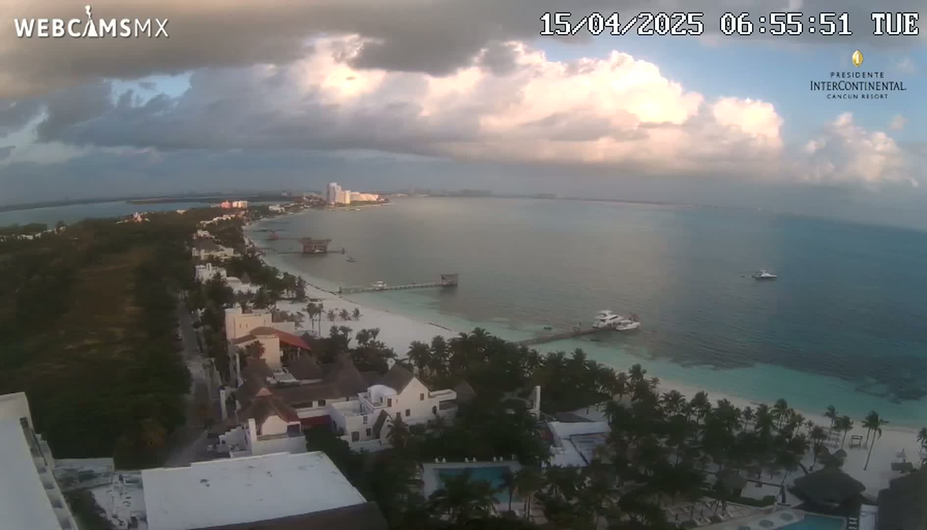 An aerial view of a tropical beach with cloudy skies shows a coastline with hotels, palm trees, and calm ocean water.