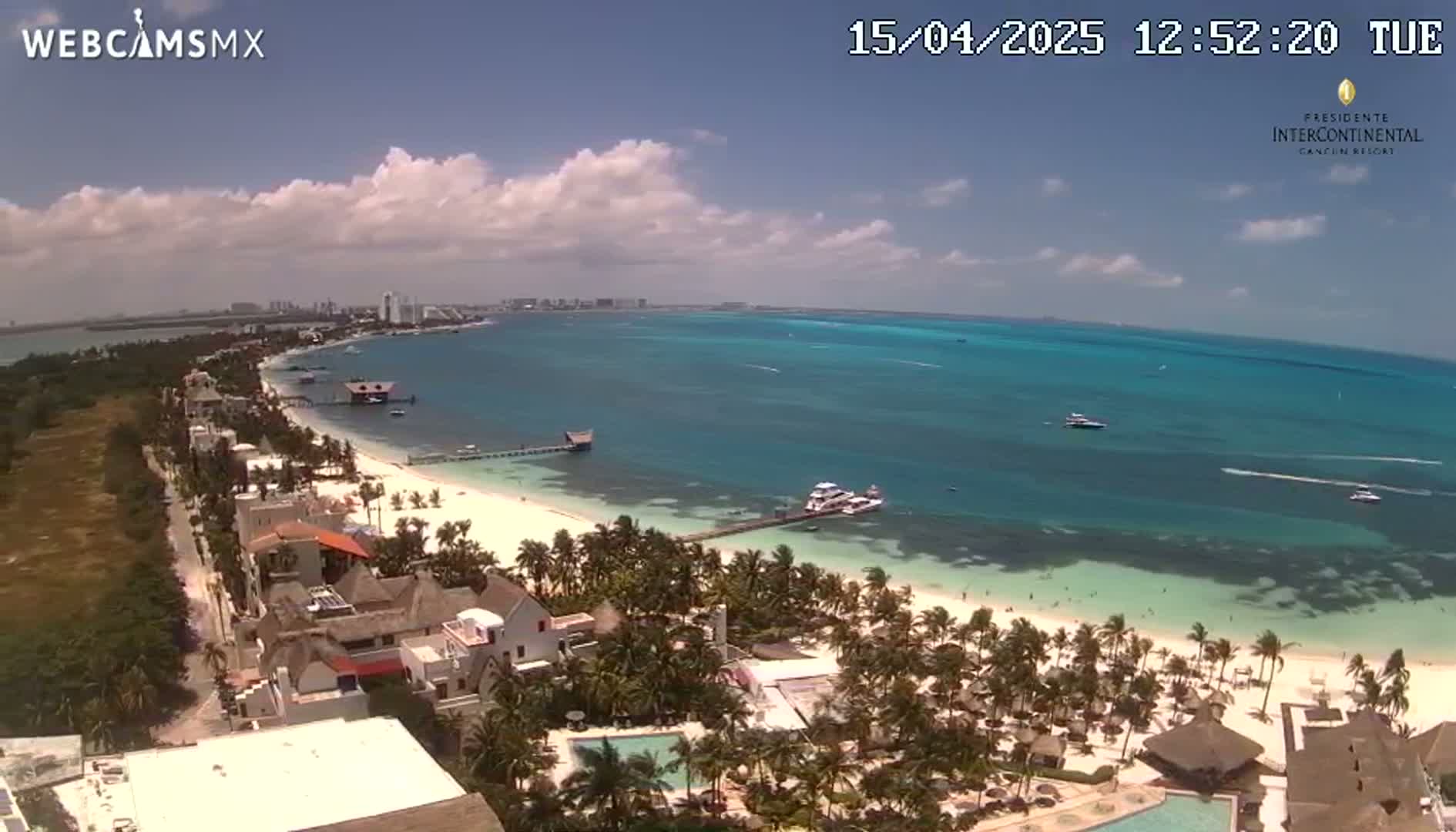 An aerial view of a tropical beach with turquoise water, under partly cloudy skies, shows a resort and a town along the coast.