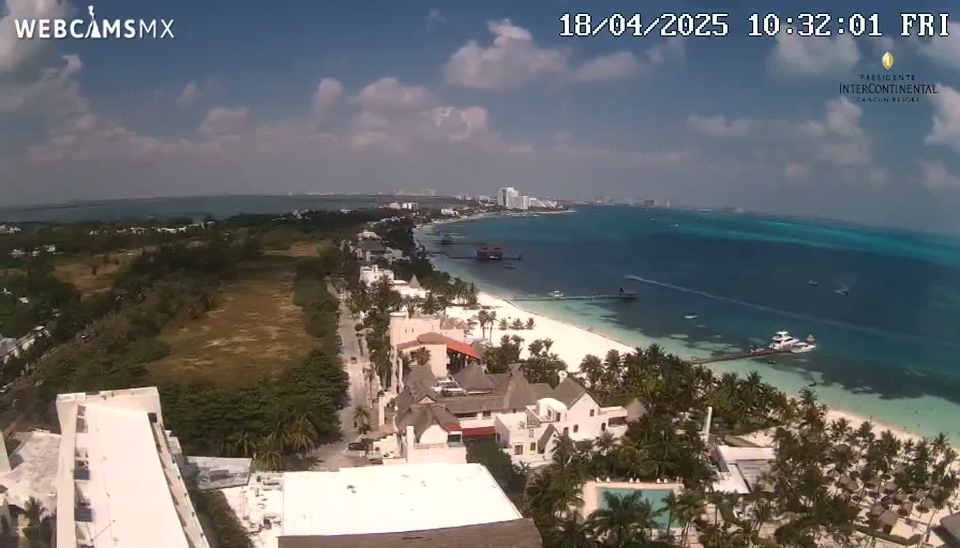 An aerial view shows a partly sunny day over a white sand beach, turquoise ocean, beachfront hotels, and lush vegetation.