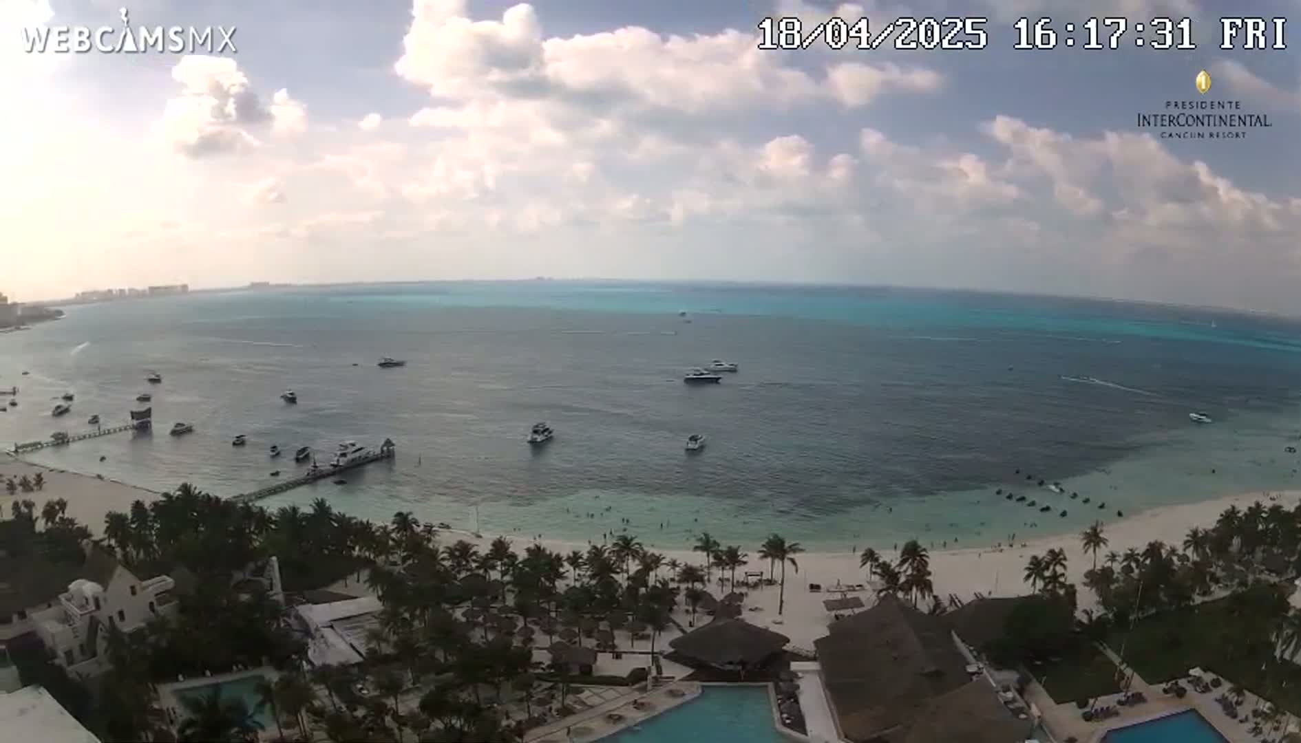 A partly cloudy day shows a tranquil beach scene with turquoise water, several boats, and a resort with palm trees and a swimming pool.