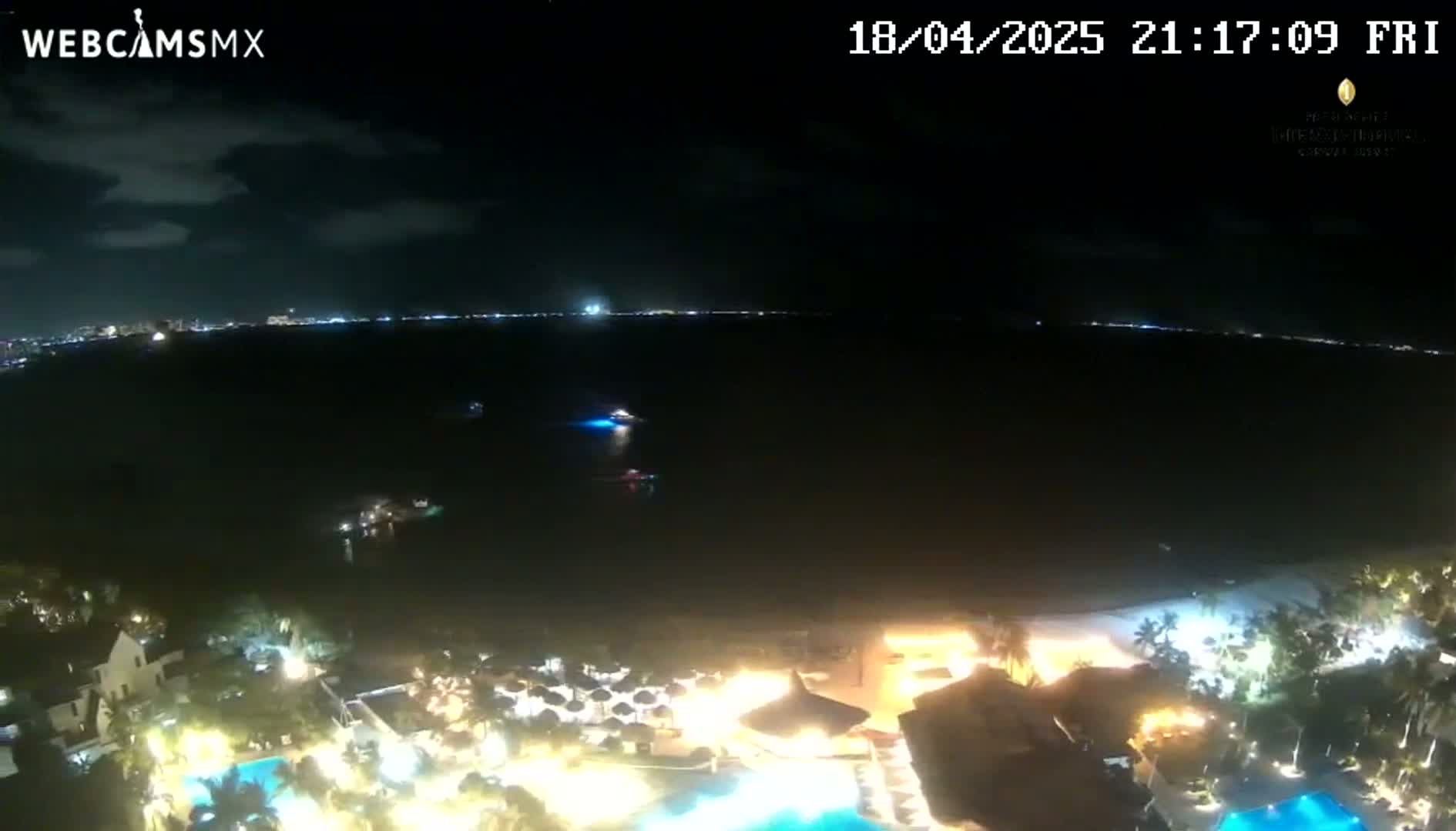 A nighttime view of a resort town on a dark, calm body of water under a partly cloudy sky.
