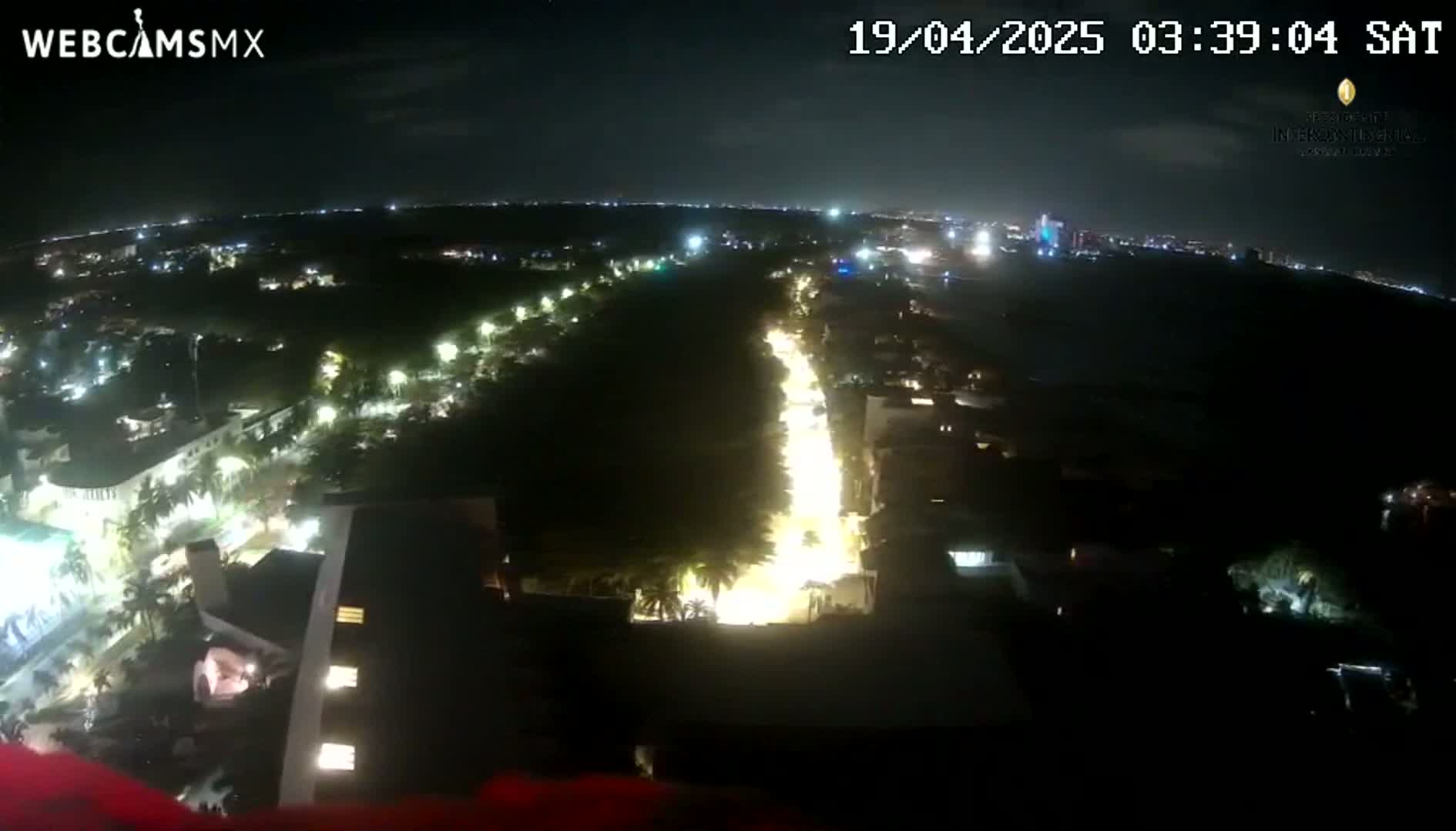 A nighttime, high-angle view shows a brightly lit city street curving through a darker, tree-lined area, with other city lights visible in the distance.