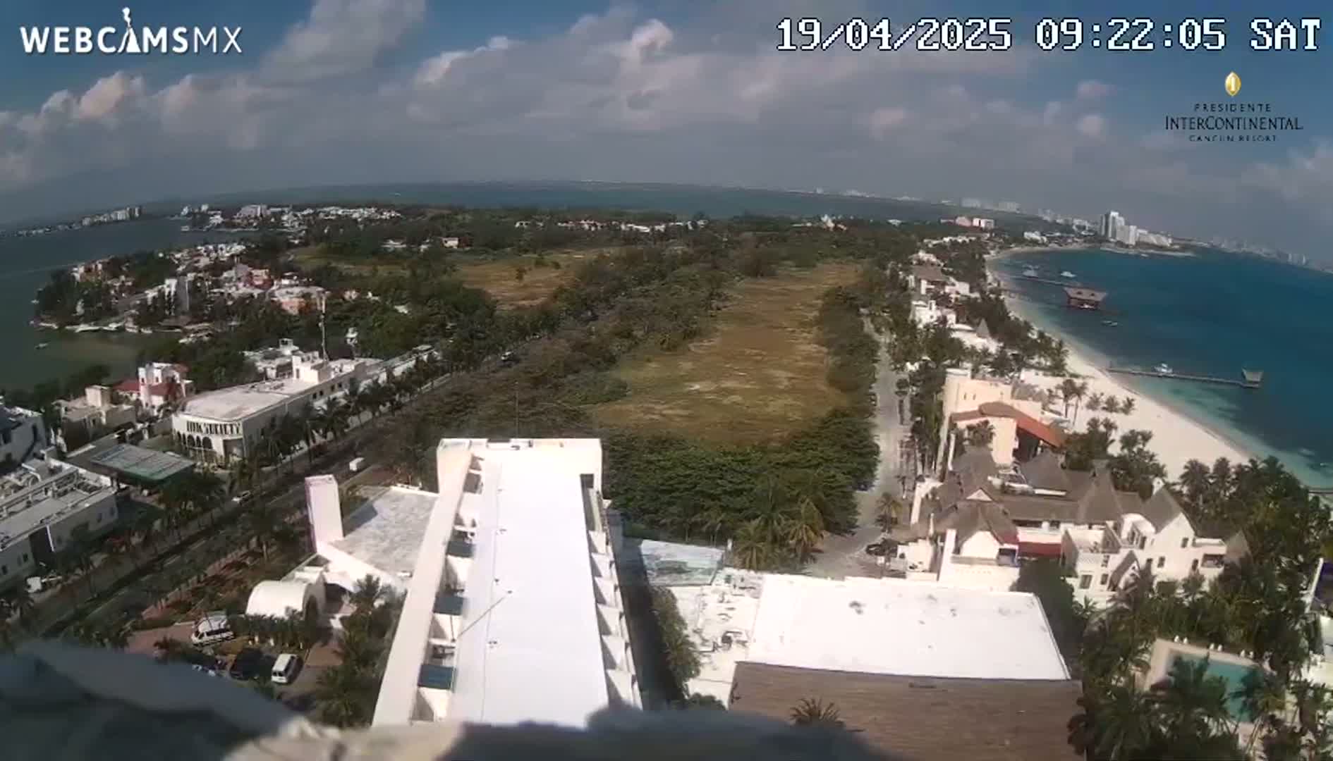 An aerial view shows a coastal area with buildings, a sandy beach, and a partly cloudy sky.