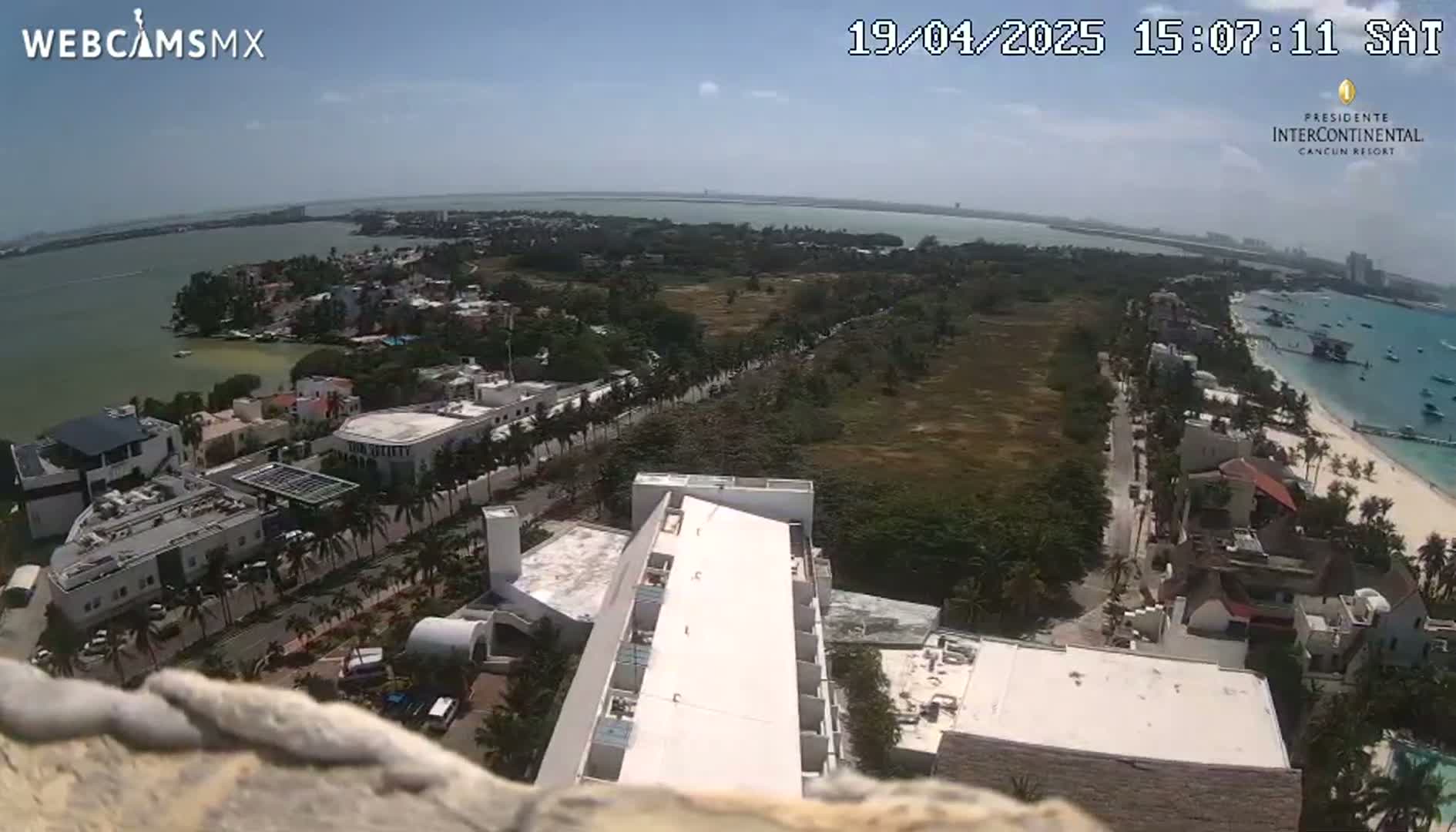 An aerial view shows a coastal town on a sunny day, with a body of water visible in the background and a white-roofed building in the foreground.