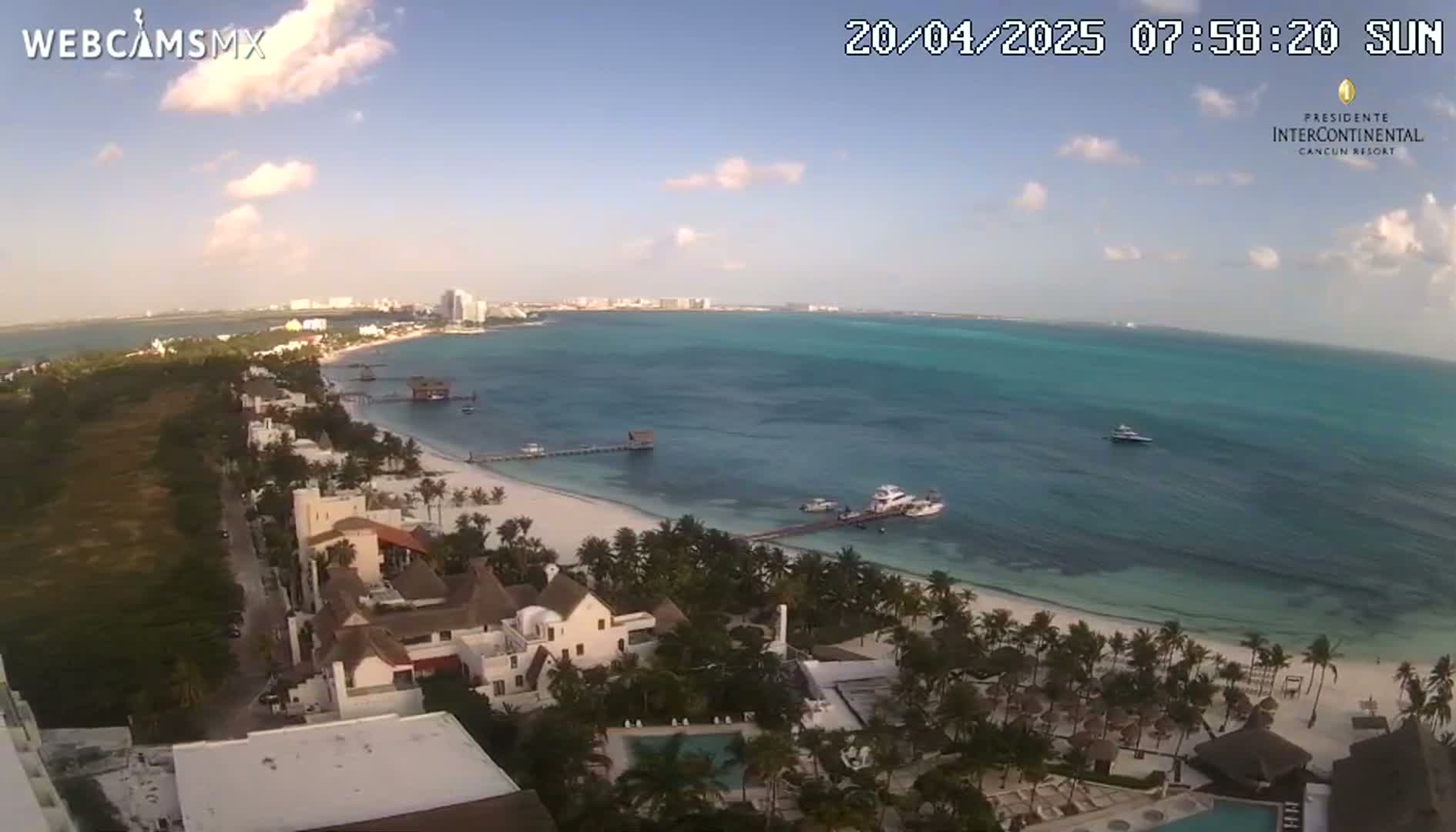 A partly sunny day reveals a white sand beach lined with palm trees and beachfront hotels, overlooking a calm turquoise ocean with a few boats.
