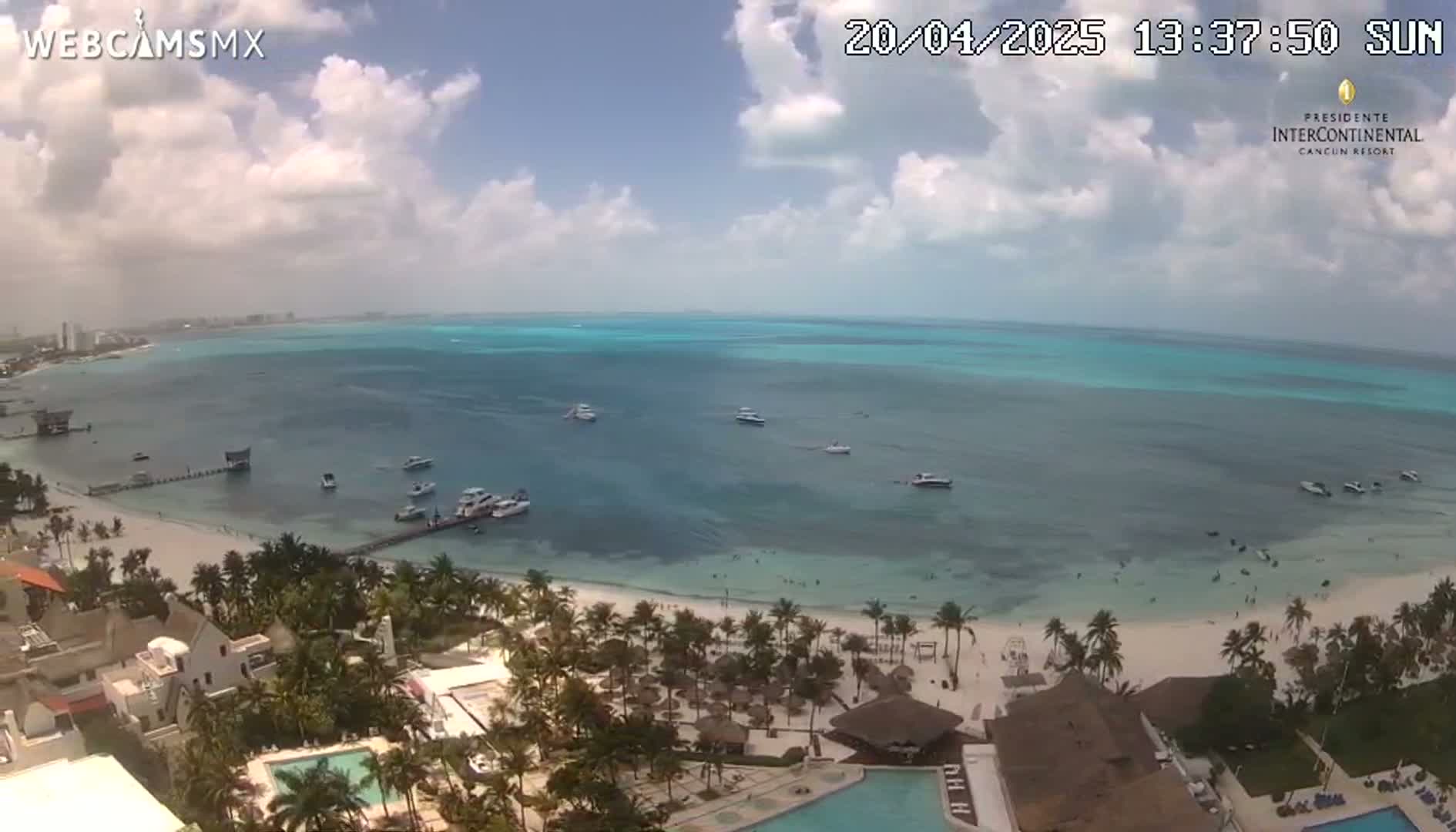 A partly cloudy sunny day shows a beach with turquoise water, several boats, and a resort with swimming pools.