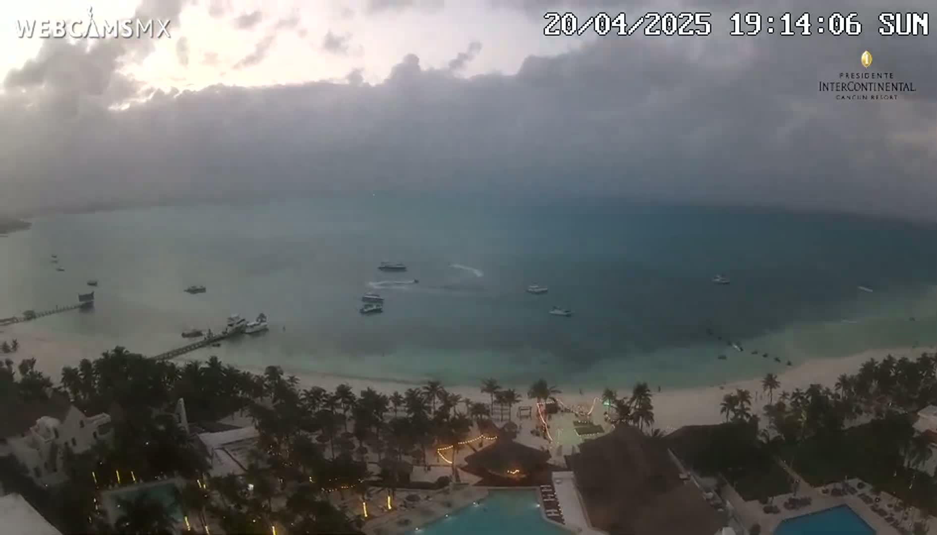 An elevated view shows a beach resort at dusk under an overcast sky, with calm ocean water and several boats visible offshore.