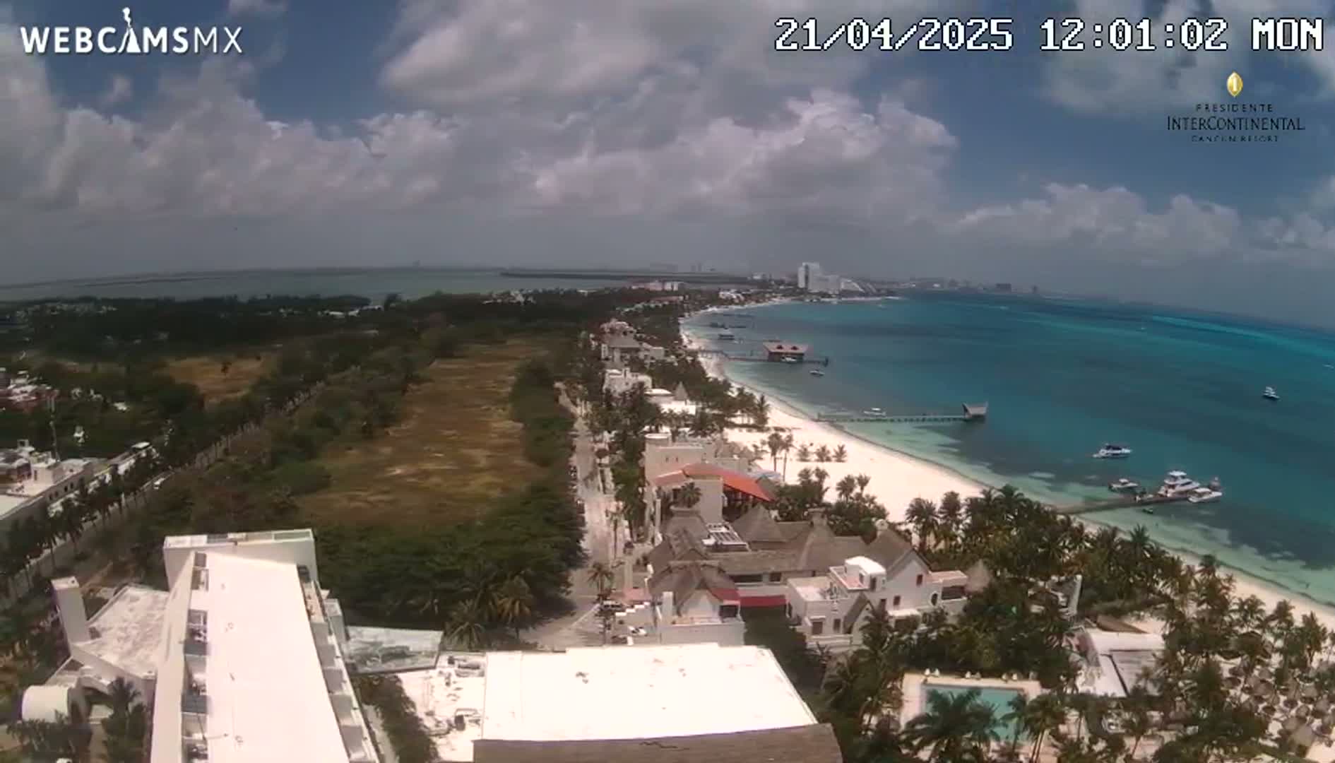 An aerial view shows a partly cloudy day over a tropical beach town with a white sand beach, turquoise water, and buildings along the coast.