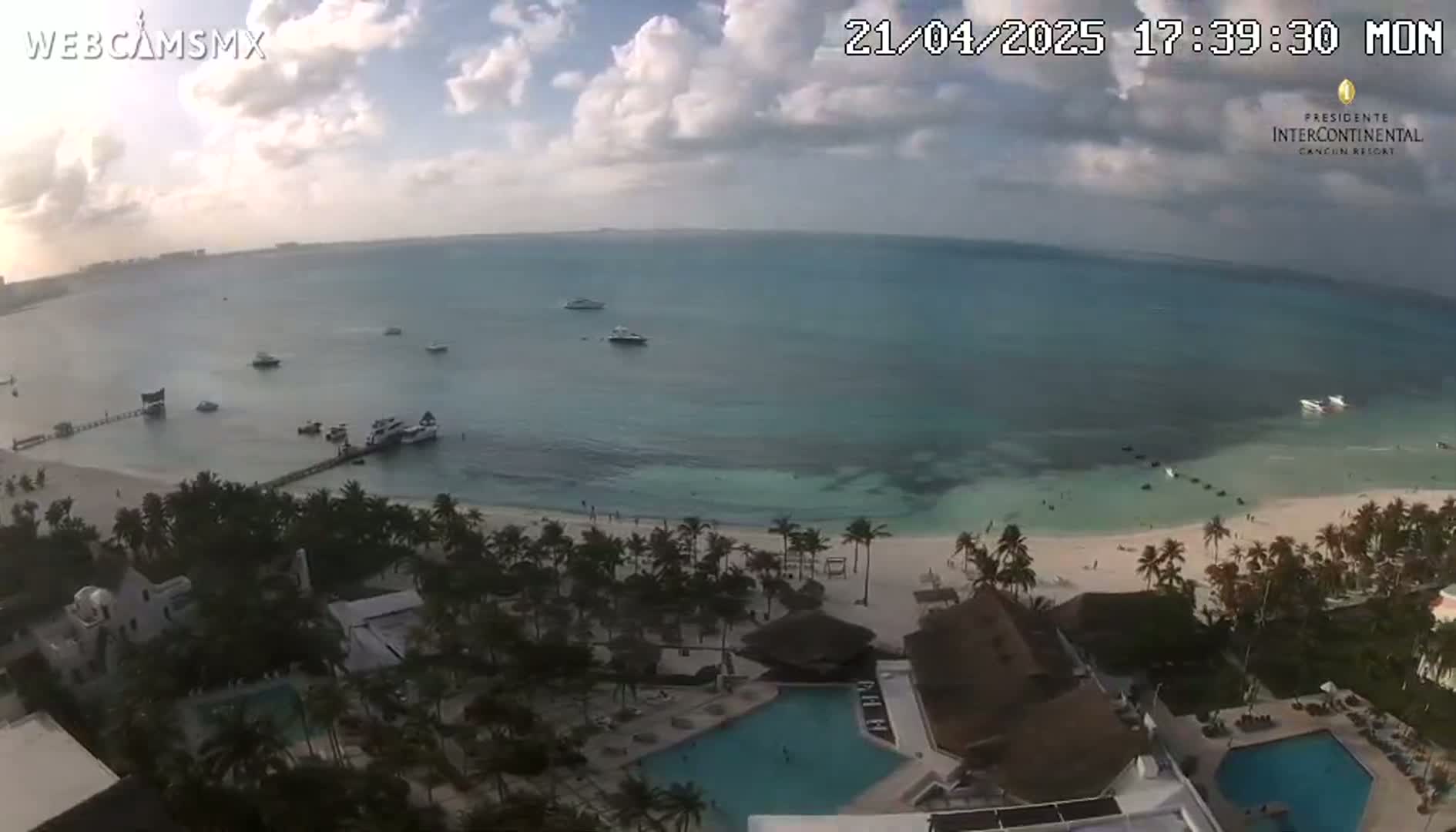 An aerial view shows a partly cloudy day at a tropical beach resort with a calm turquoise ocean, several boats, and multiple swimming pools.