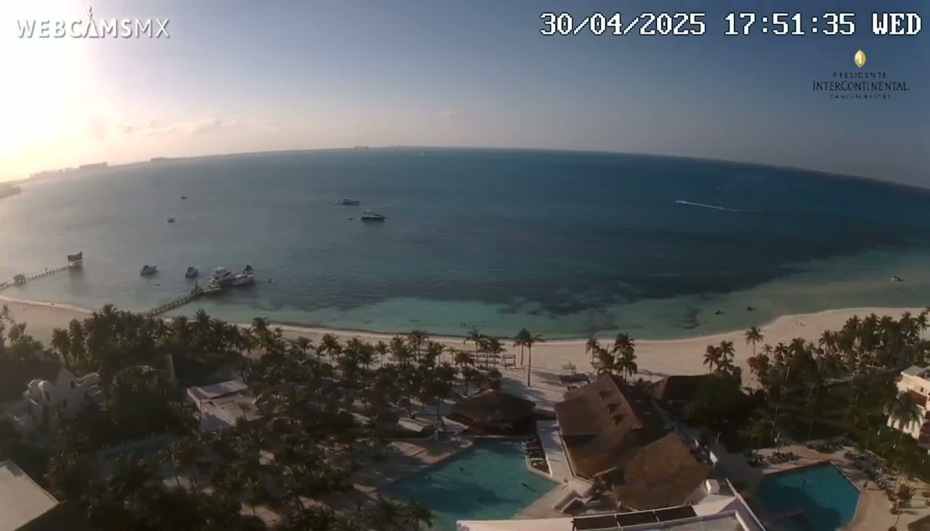 An aerial view shows a tropical resort with a swimming pool, bordering a white sand beach and calm turquoise ocean water under a clear sky.