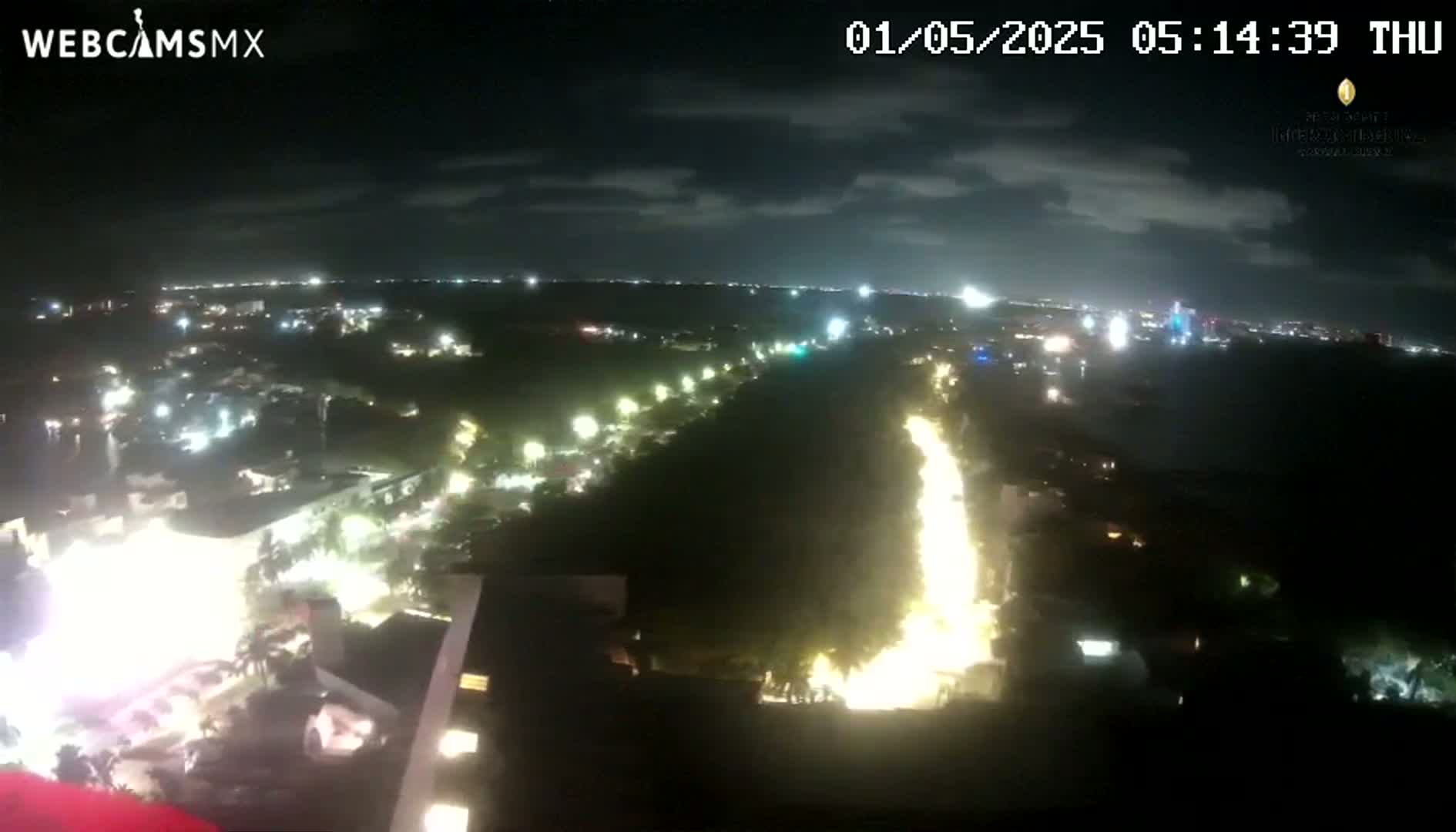 A nighttime aerial view shows a brightly lit road winding through a dark, wooded area, with other lights visible in the distance under a partly cloudy sky.