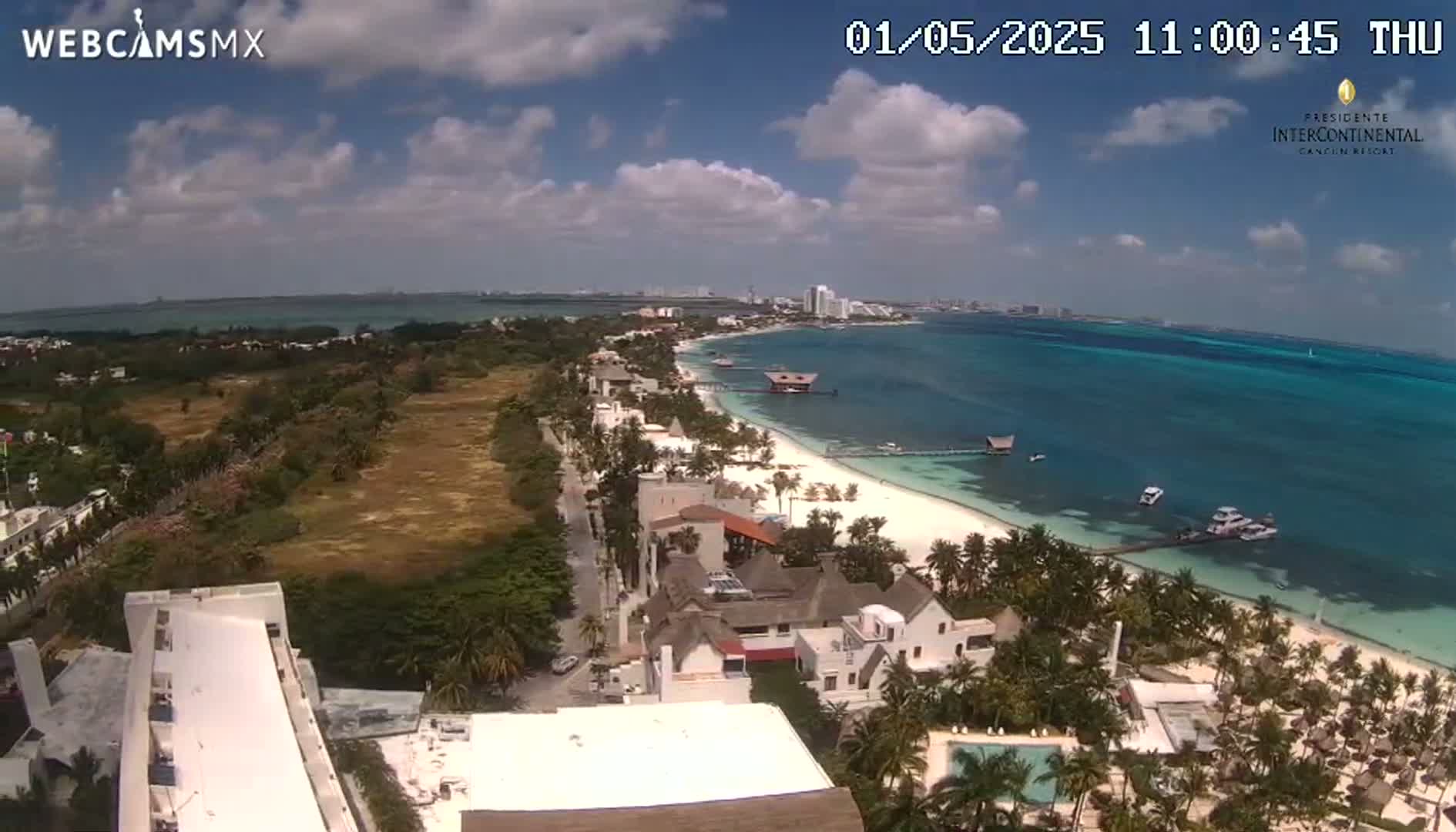 An aerial view shows a partly sunny day overlooking a beach with turquoise water, palm trees, and beachfront buildings.