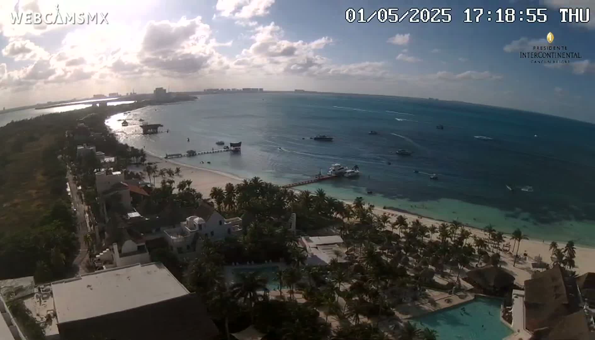 An aerial view shows a tropical beach with calm, clear water, boats on the ocean, palm trees, and resort buildings under a partly sunny sky.