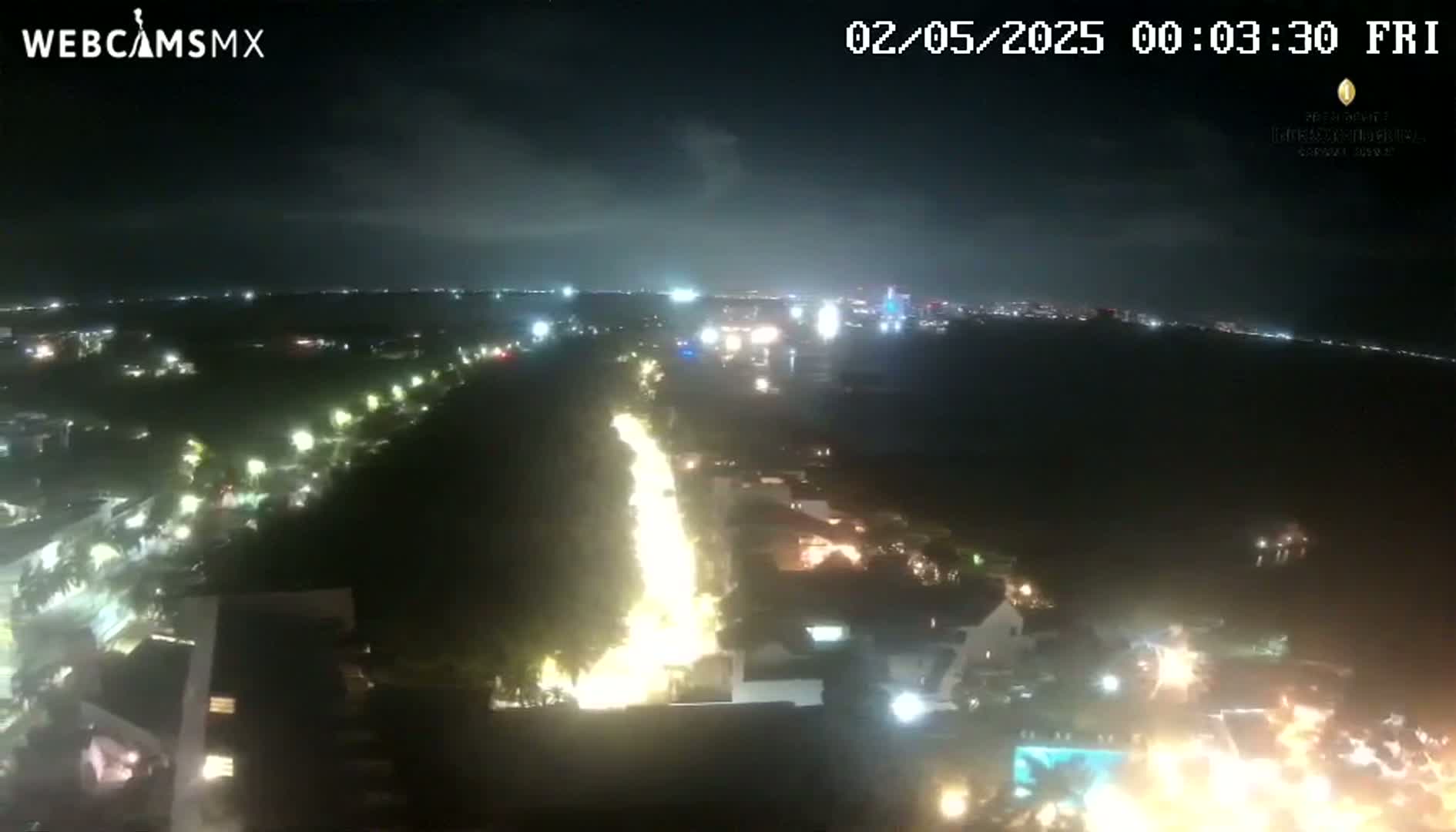 A nighttime, high-angle view of a brightly lit city street and surrounding buildings, extending to a dark coastline under a partly cloudy sky.