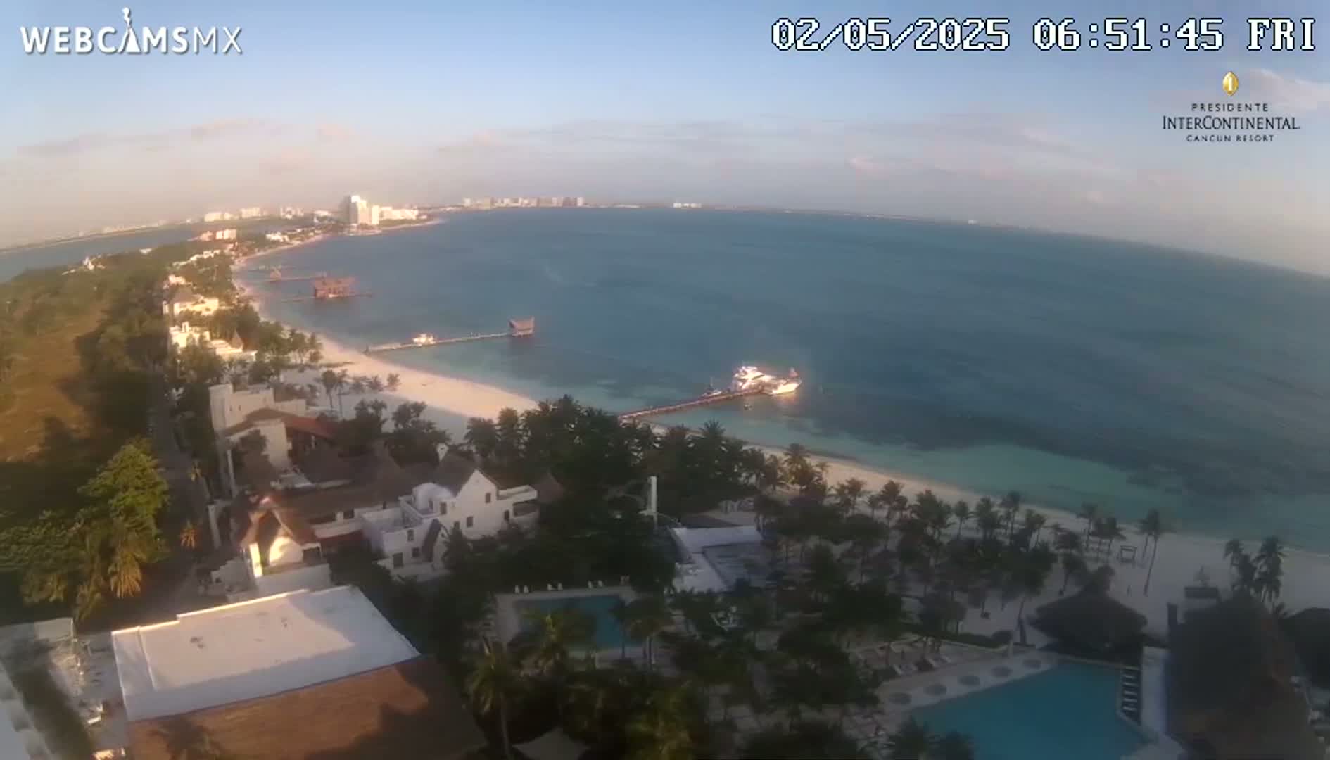An aerial view shows a beach with calm, clear water, buildings, palm trees, and pools under a clear, bright sky.