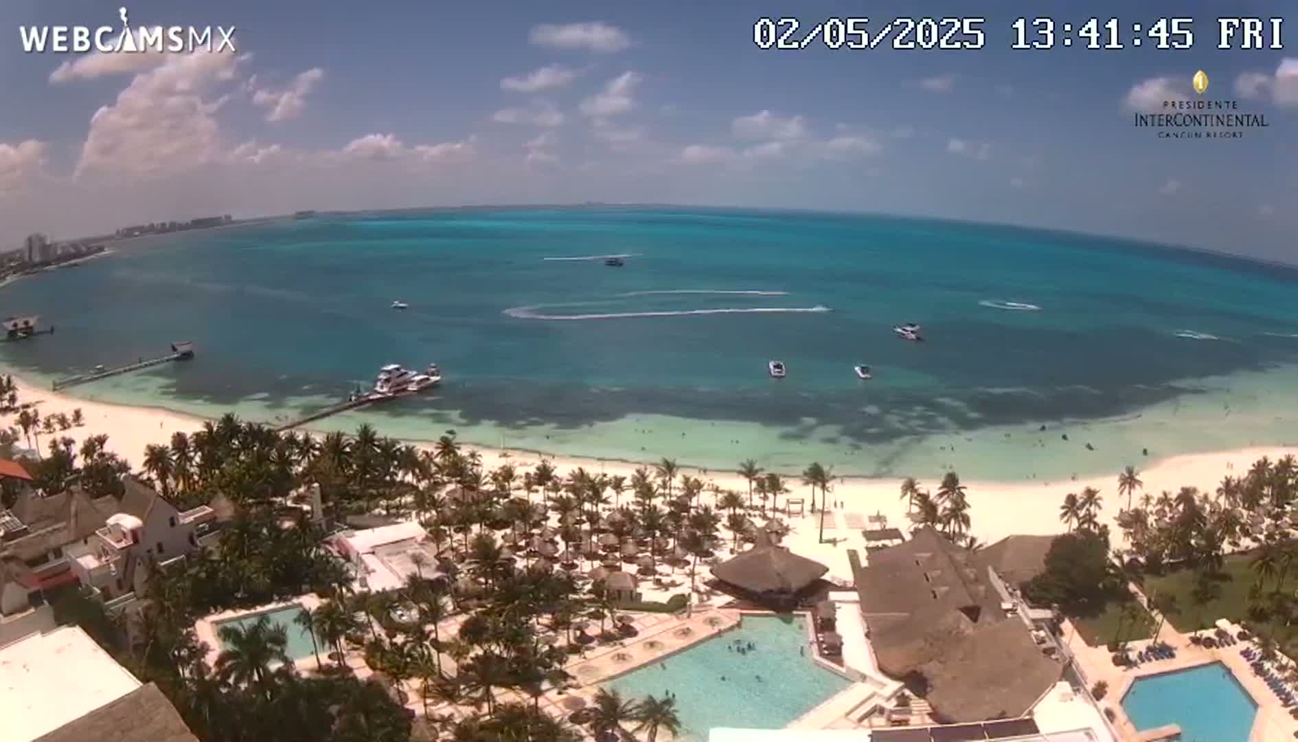 An aerial view shows a tropical beach with turquoise water, palm trees, and resort buildings under a mostly sunny sky.