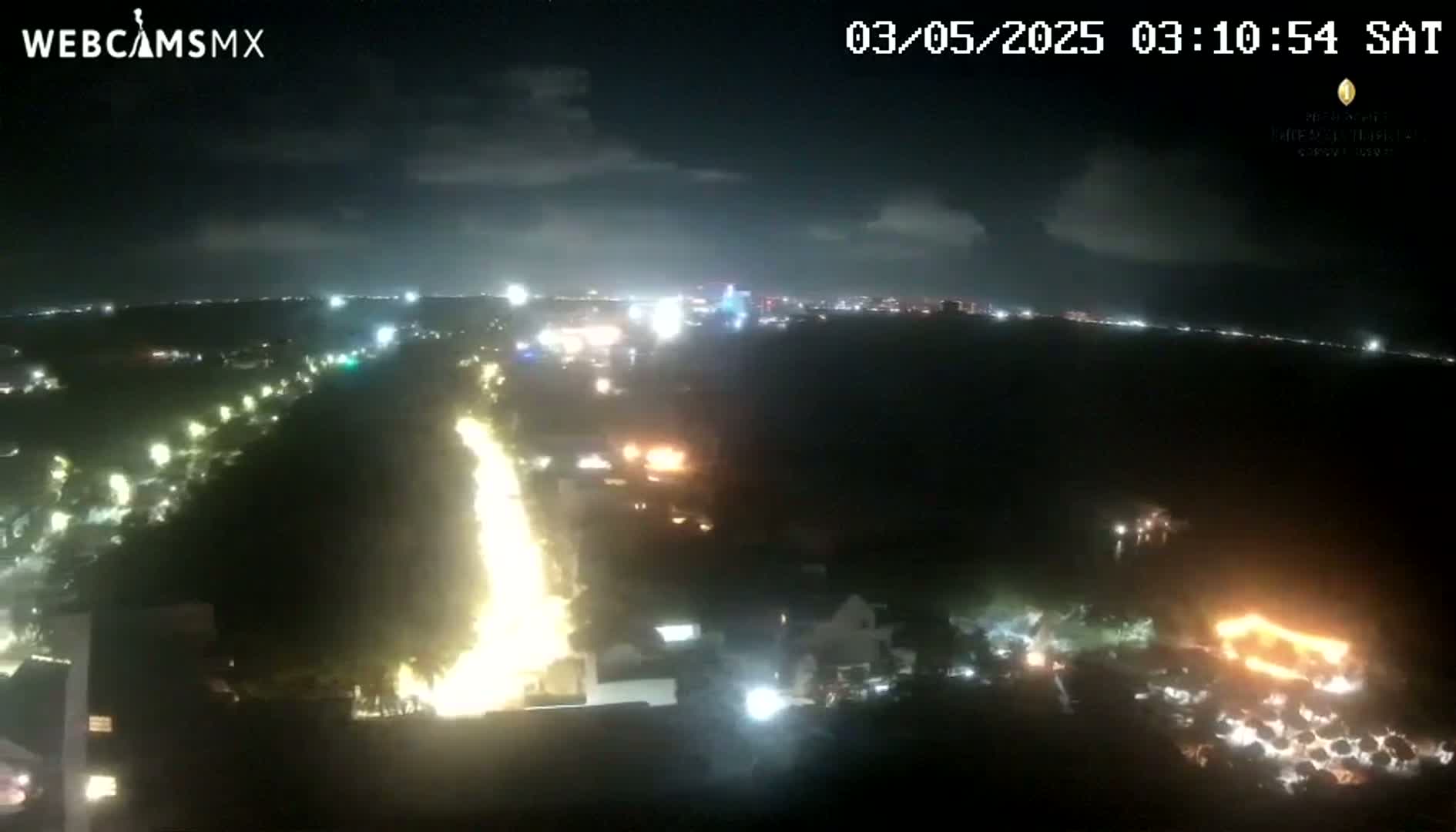 A nighttime aerial view shows a long, bright line of lights along a road, with other lights scattered across a dark landscape under a partly cloudy sky.