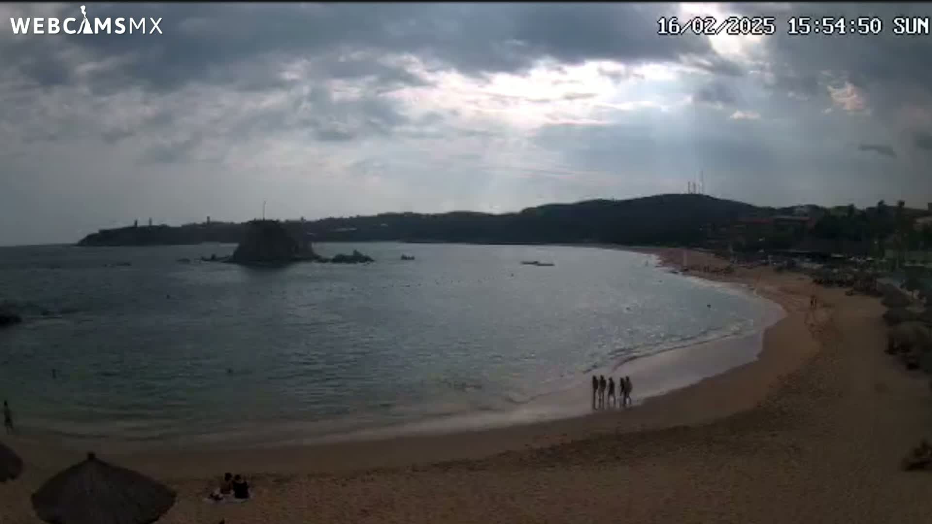 A sandy beach curves into a calm bay under a partly cloudy sky, with a few people visible on the sand and in the water.