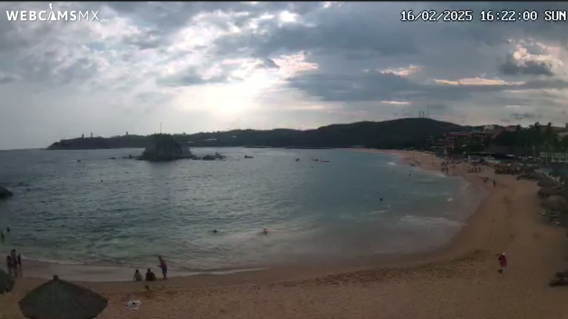 A sandy beach with calm water and a few people on it is partially visible under a cloudy sky.