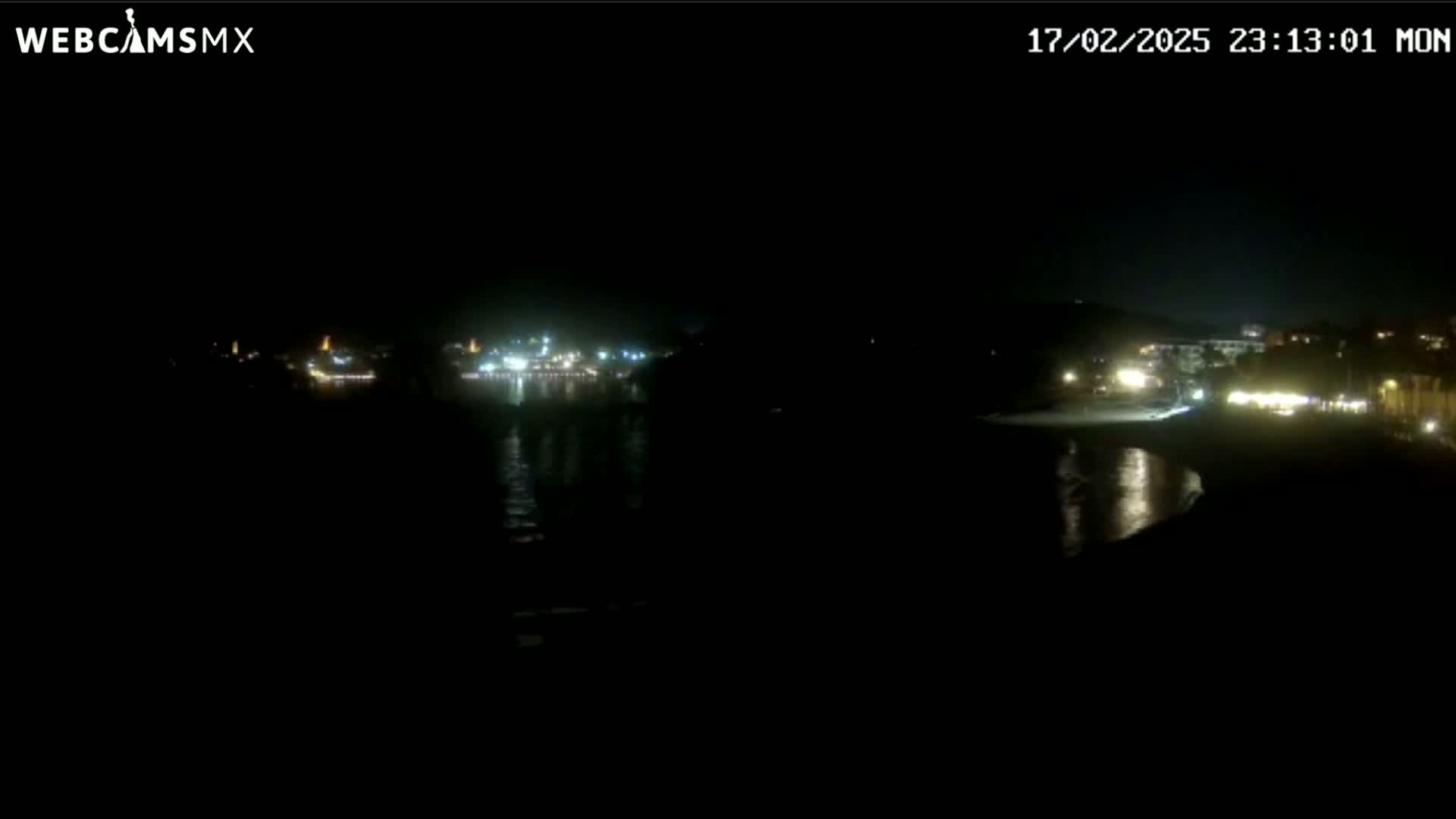 A nighttime view of a dark coastline with dimly lit buildings and a calm body of water reflecting some lights.