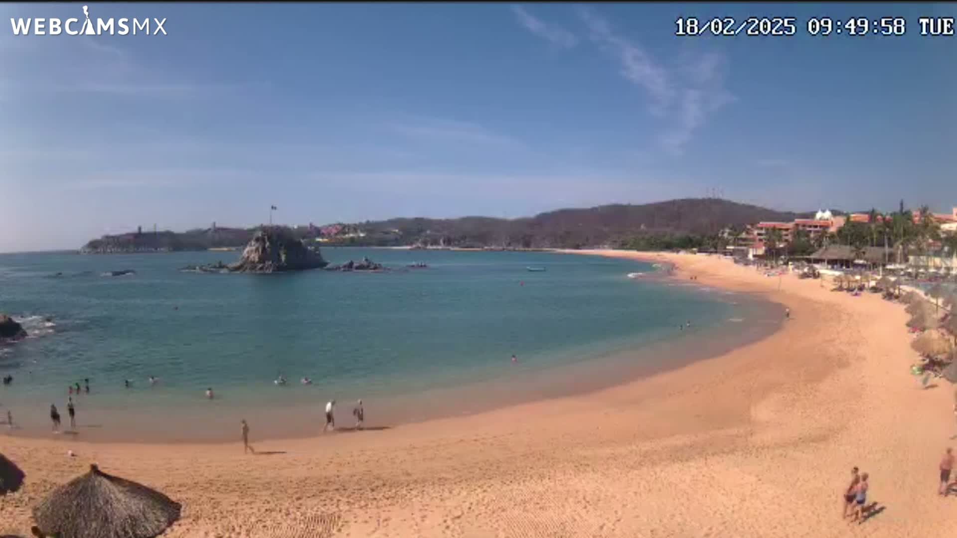 A sandy beach curves around a calm, clear bay under a sunny sky, with people swimming and relaxing on the shore and a small island visible in the distance.