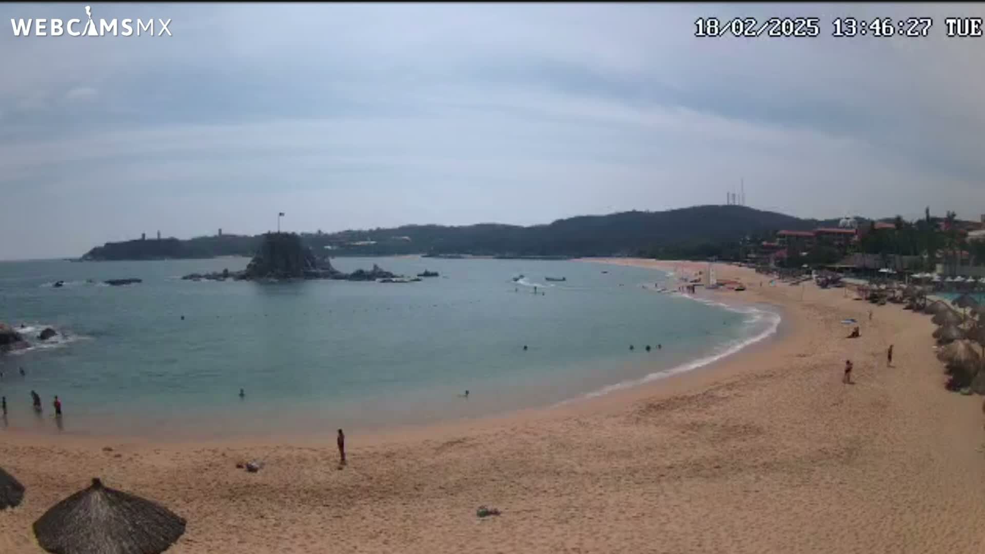 A mostly sunny day at a secluded beach cove with calm, clear water, featuring a rocky island, a sandy shore with scattered people, and buildings visible in the distance.