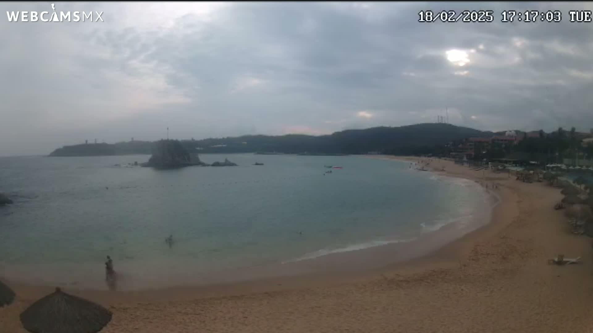 A sandy beach curves around a calm bay under a mostly cloudy sky, with a few people visible on the beach and in the water.