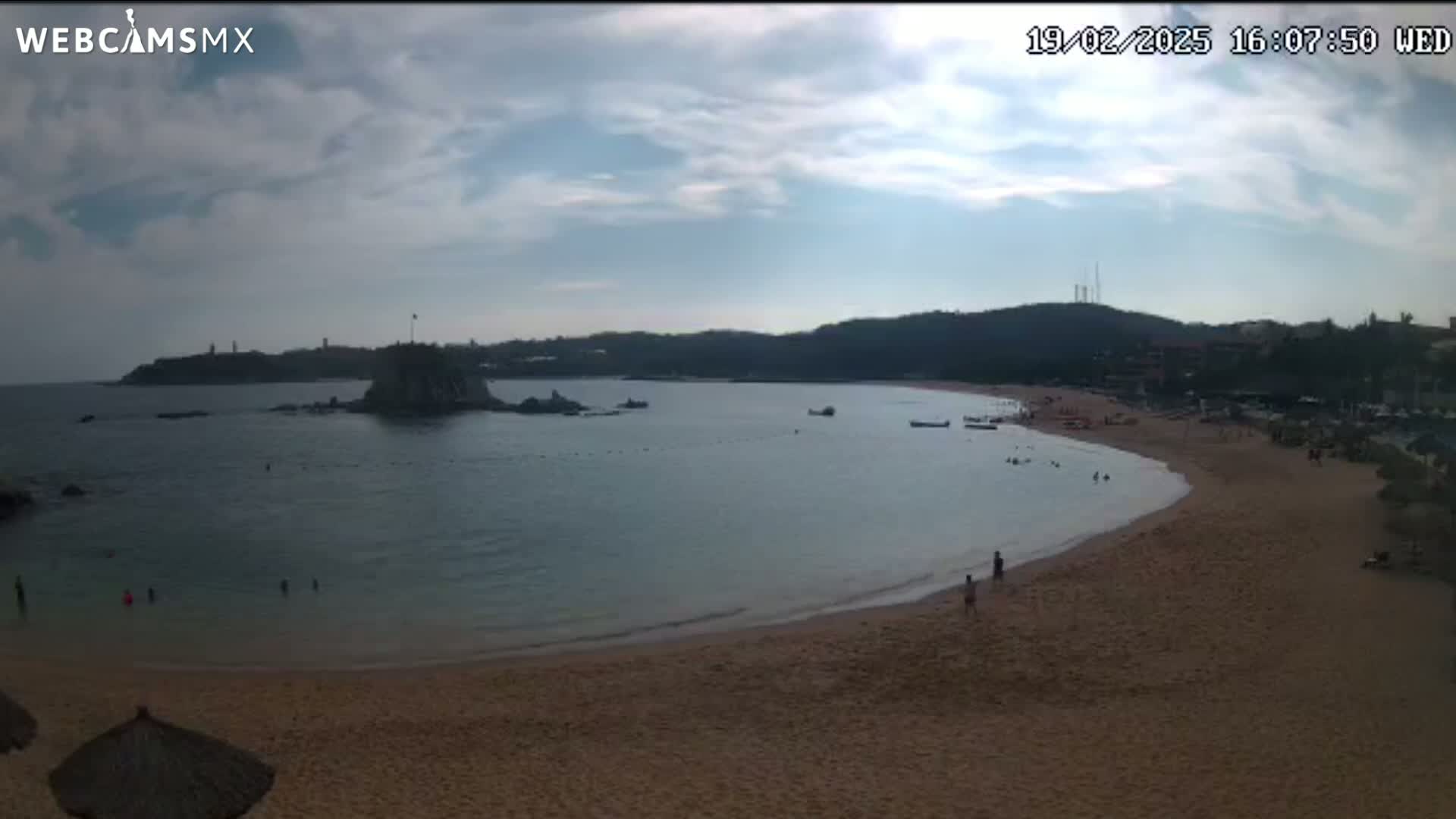 A partly cloudy day at a sandy beach cove with calm water, a few people swimming and walking on the shore, and small boats in the bay.
